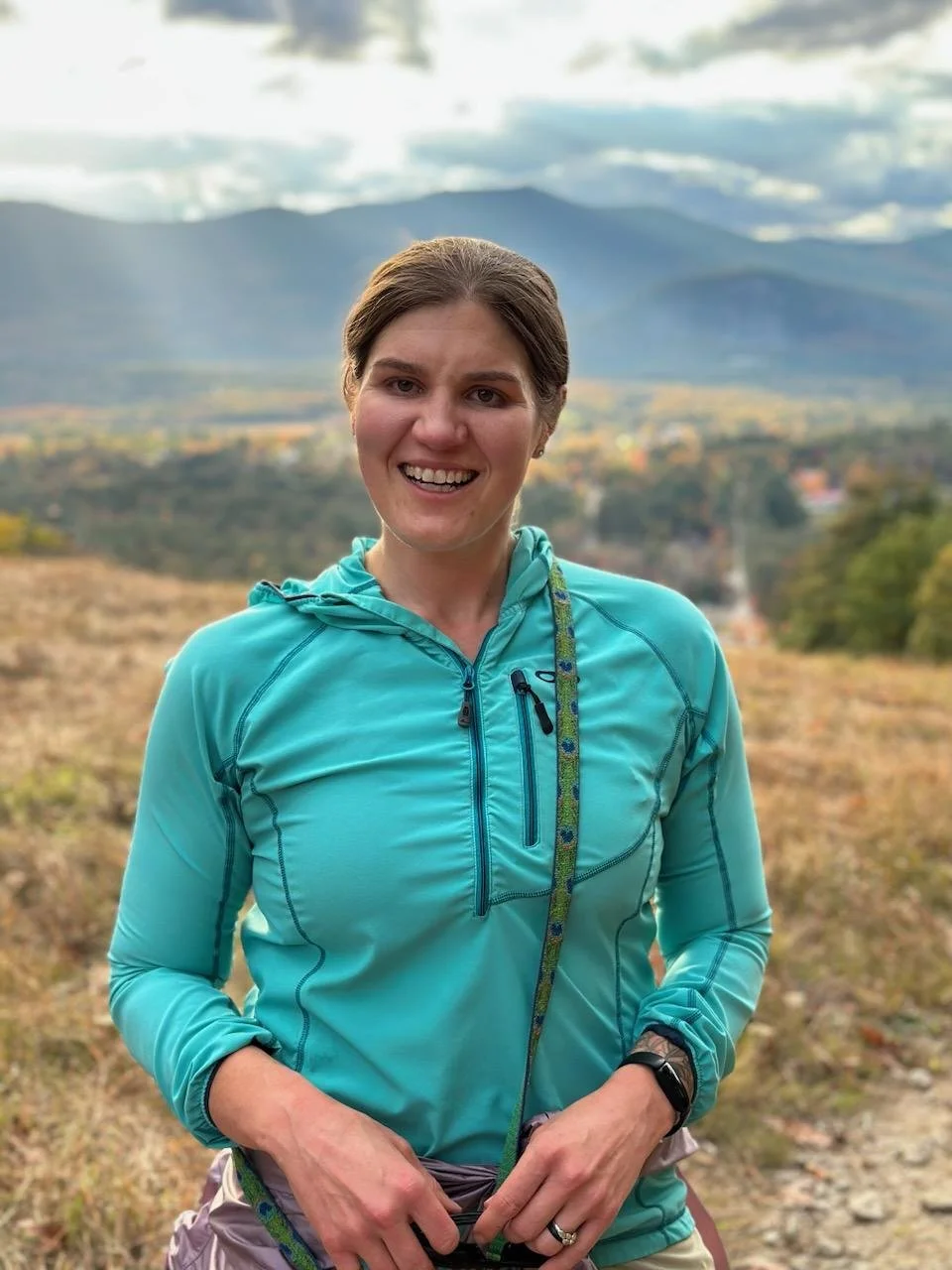 Owner Liz Prior, A woman in a turquoise outdoor jacket smiling outdoors with mountains and cloudy sky in the background.