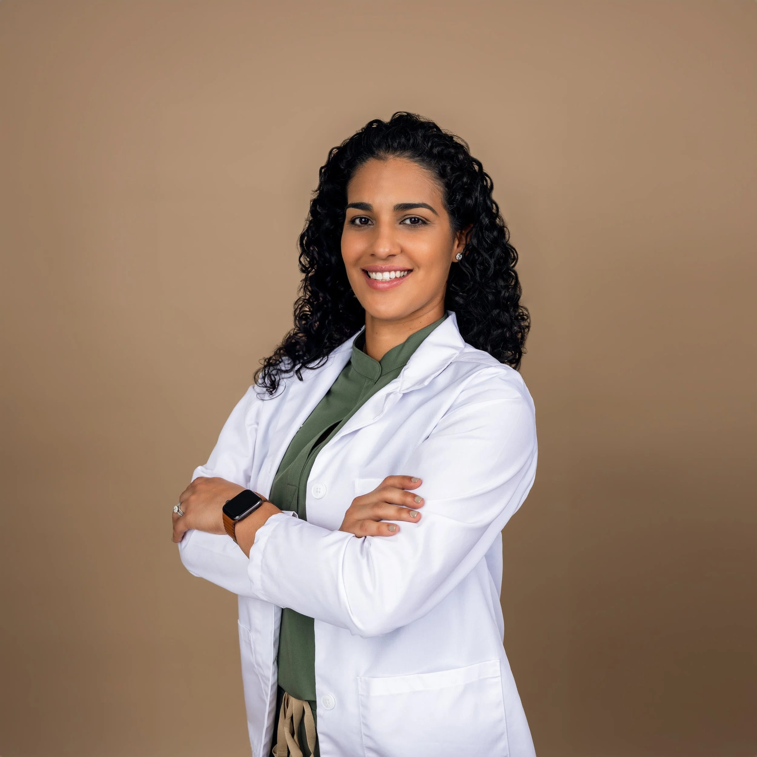 A smiling female healthcare professional with curly black hair, wearing a white lab coat over a green top, standing with arms crossed against a neutral background.