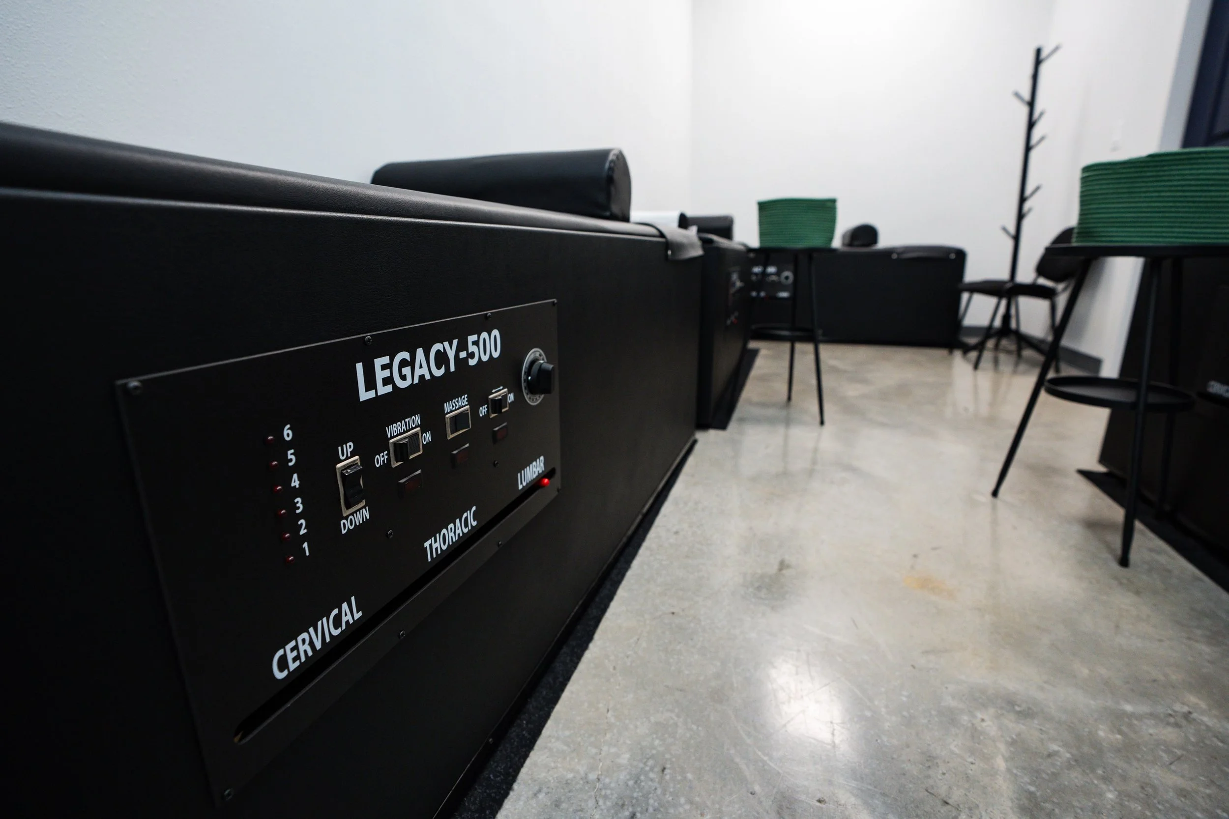Black massage table with control panel labeled LEGACY-500 in a room with chairs, and a coat rack in the background.