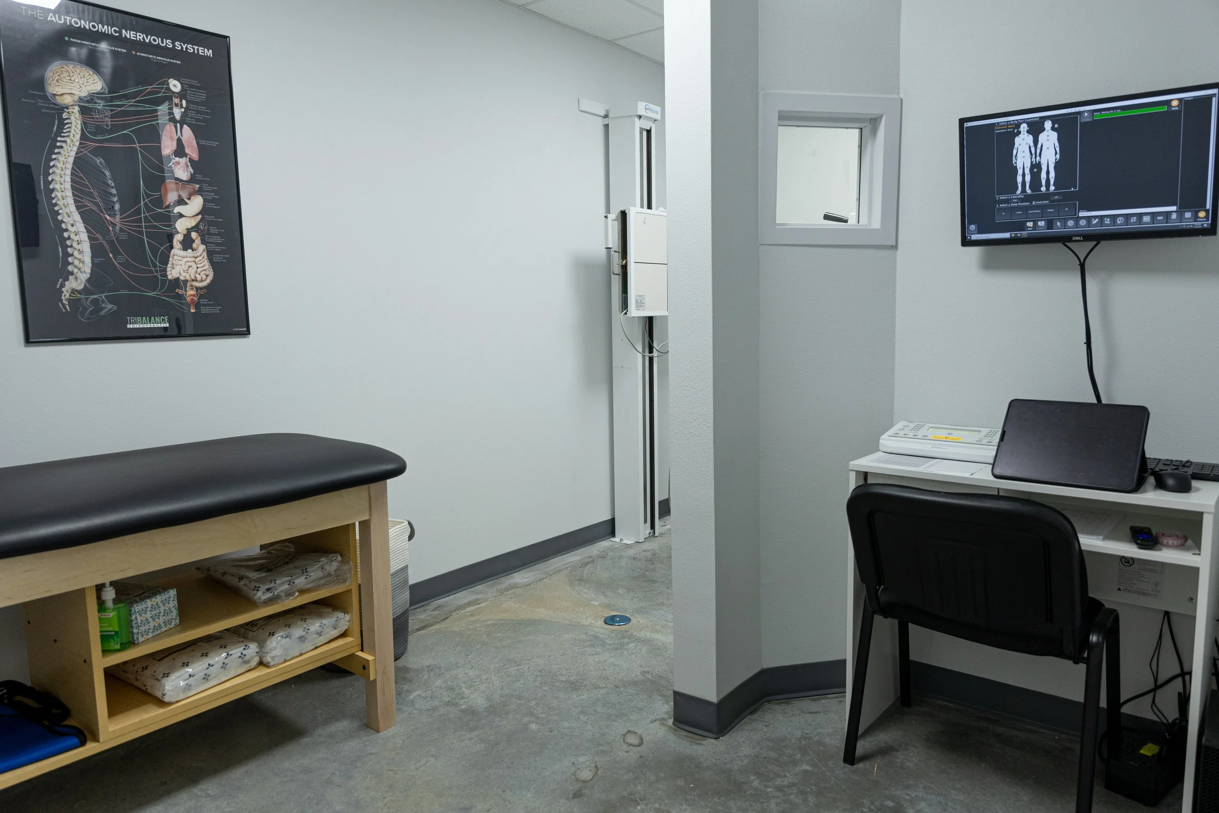 Medical examination room with an examination table, a poster of the autonomic nervous system on the wall, a computer and monitor on a white desk, and a small window.