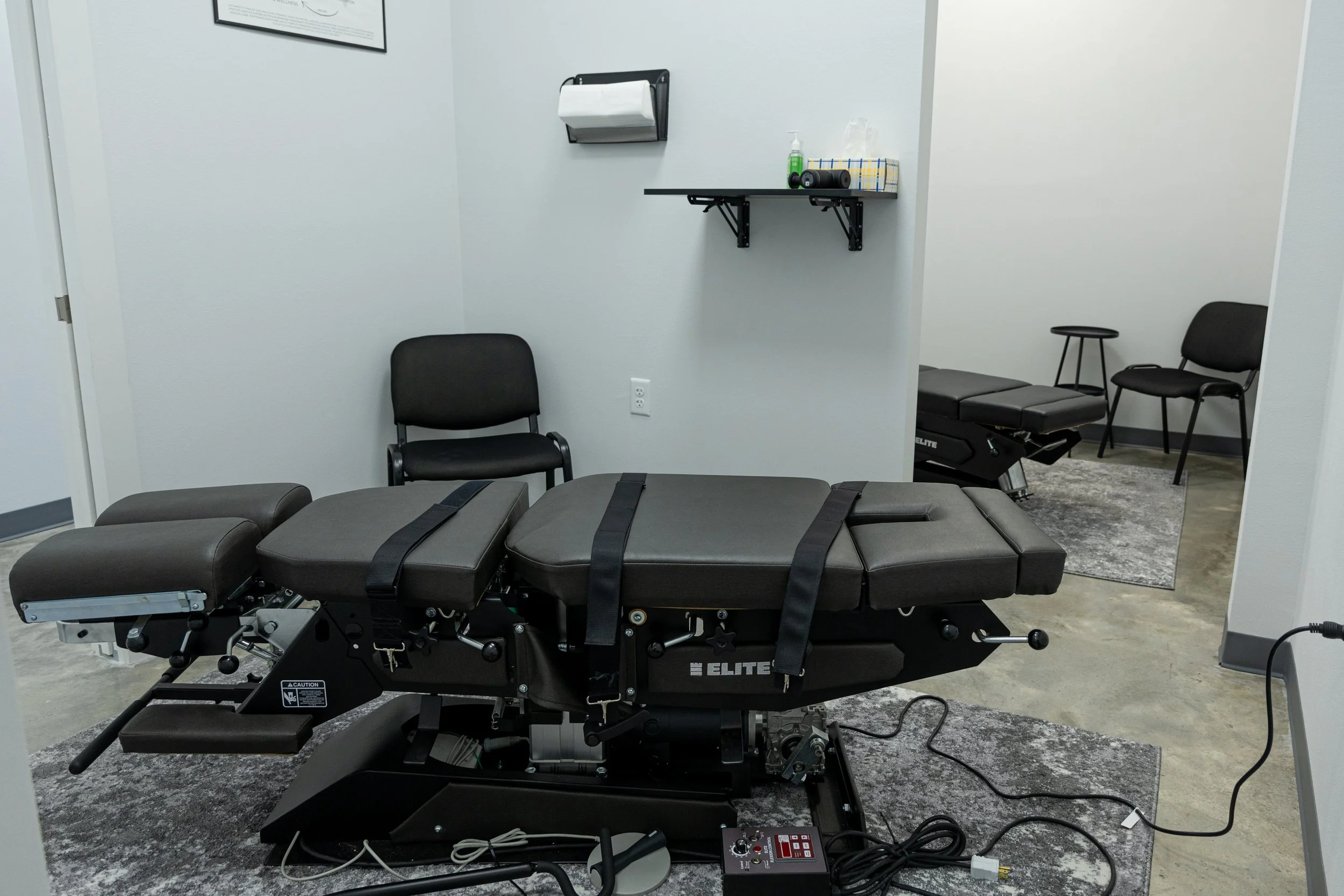Medical examination or therapy room with an adjustable treatment table, a black chair, and additional chairs in a corner. Wall-mounted shelf with supplies, and a framed certificate or artwork on the wall. The room has gray carpeting and concrete flooring.