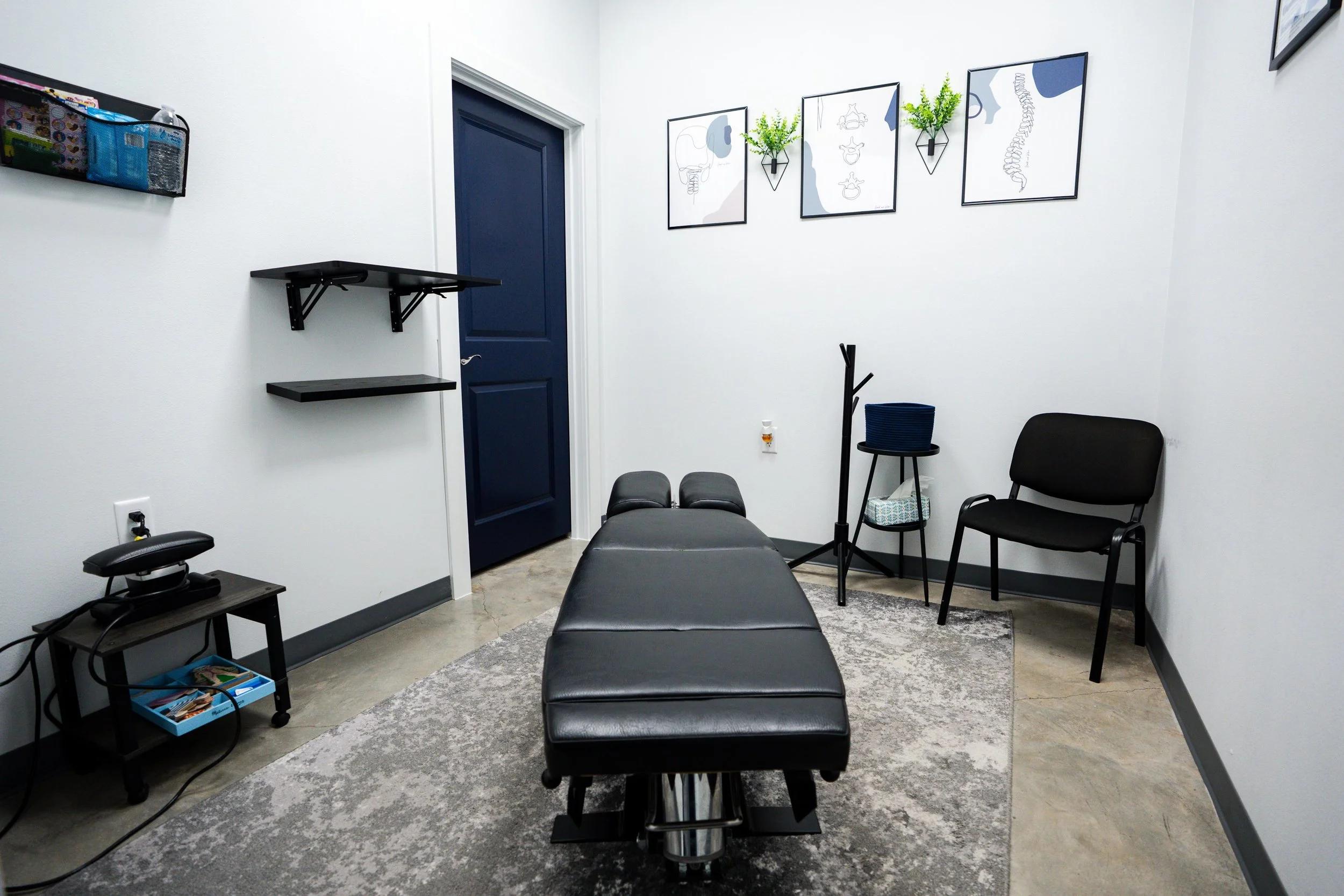 A treatment room with a massage table, chairs, wall art, and storage in a clean, minimalist space.