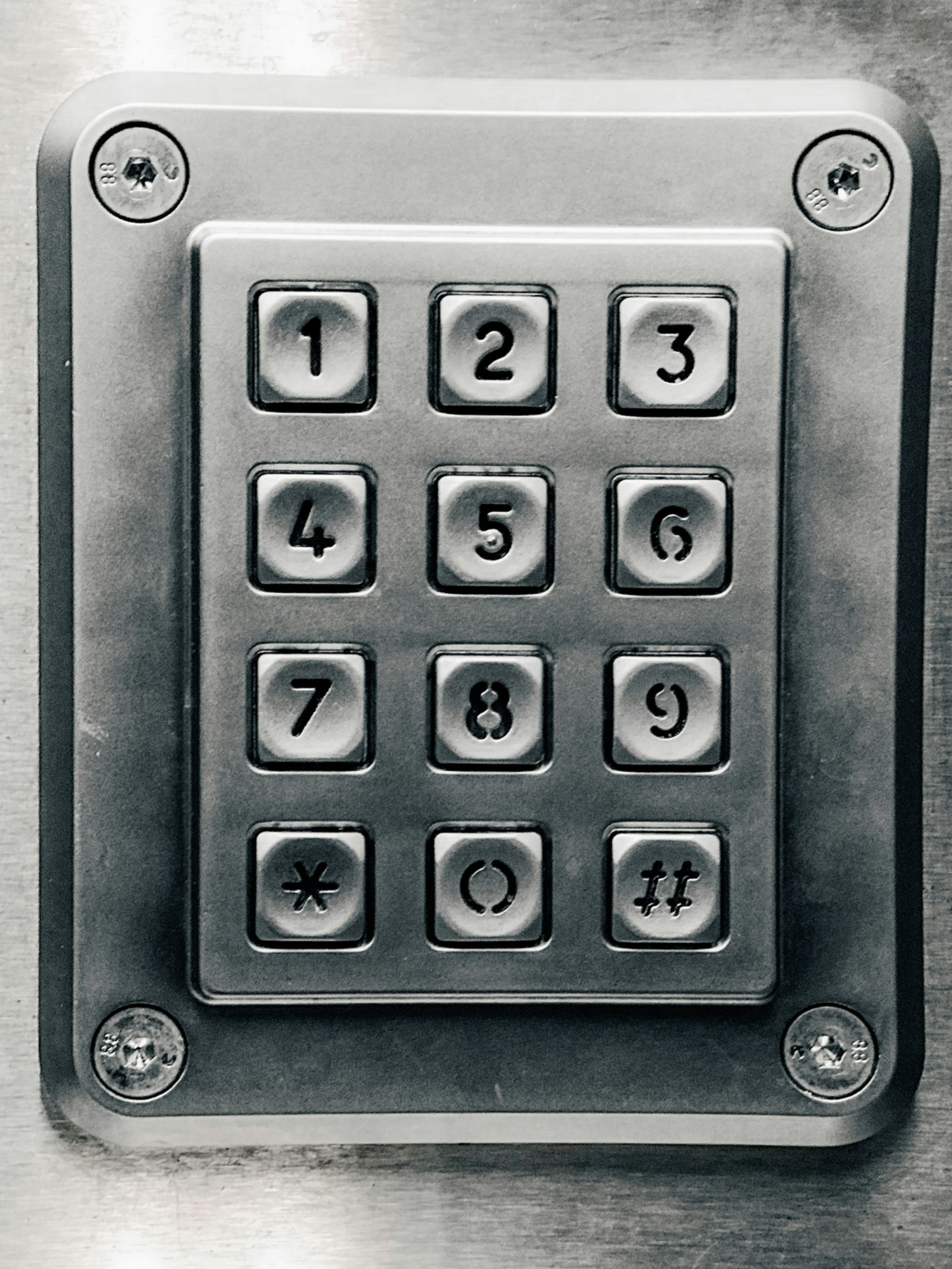A metallic keypad with numbered buttons from 1 to 9, along with an asterisk, zero, and pound/hash symbols, mounted on a flat metallic surface with four screws, one at each corner.
