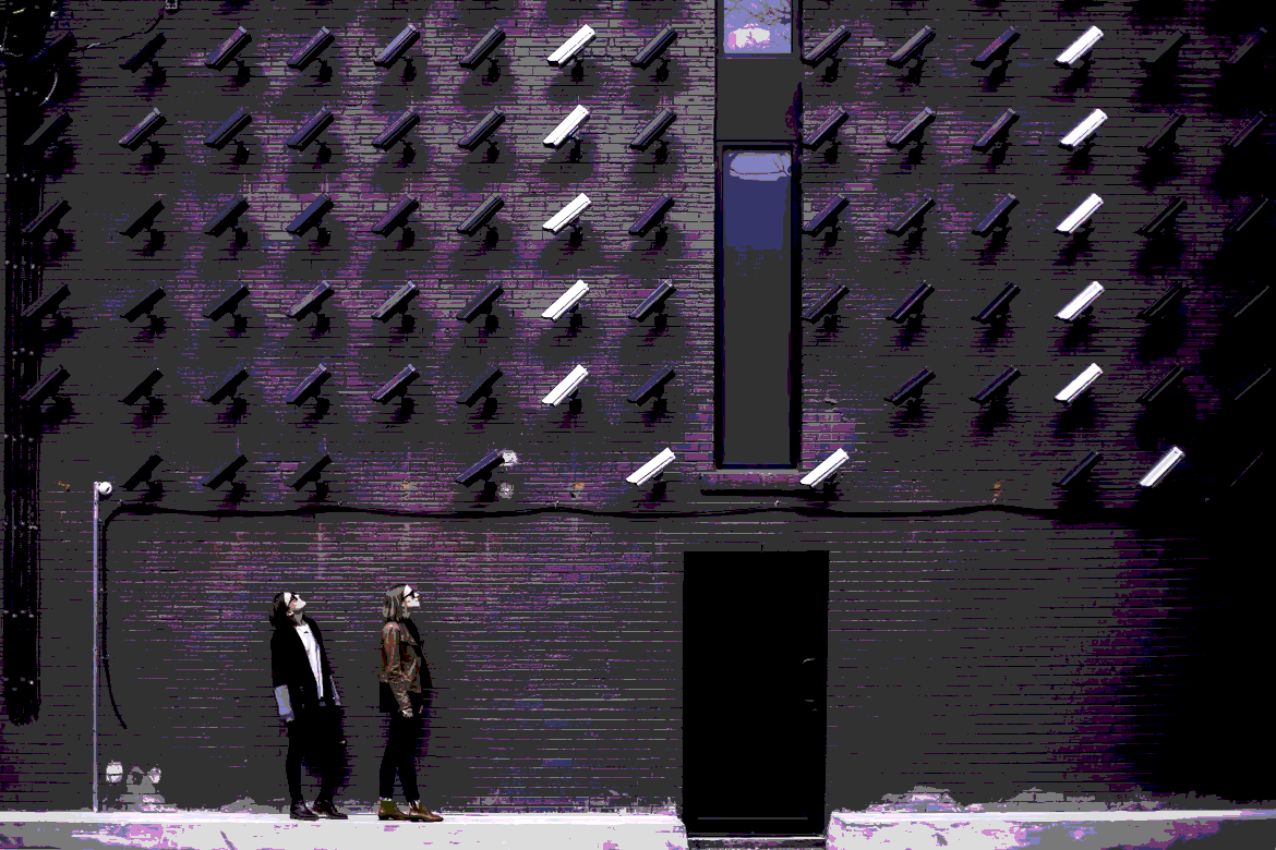 Two people walk along a sidewalk in front of a dark brick building with rows of exterior lights mounted on the wall, some turned on and some off.
