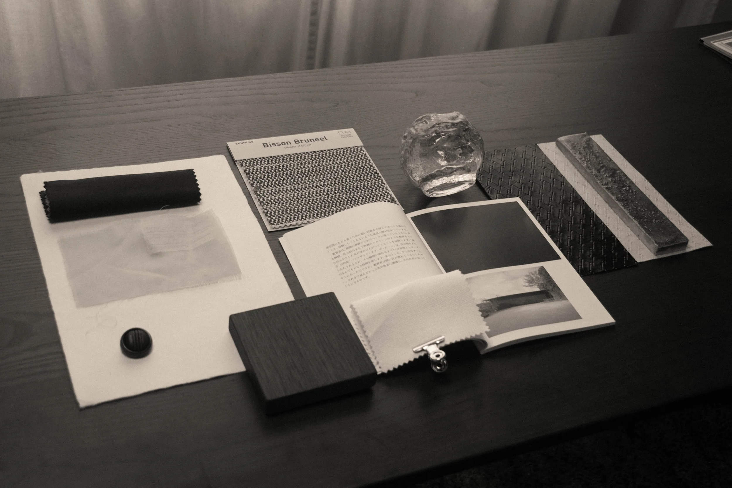 Interior material palette for a Studio Cér project, featuring fabric swatches, a glass sculptural object and stone samples on a dark wood table.