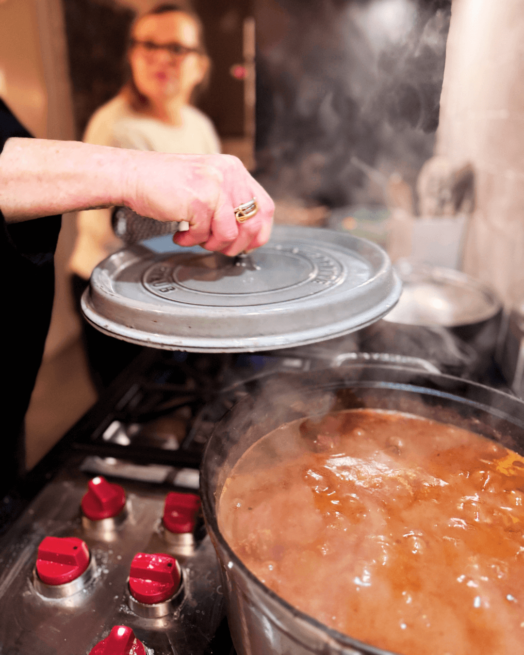 Person placing a metal lid on a pot of simmering stew on a stove, with another pot nearby, in a kitchen setting.