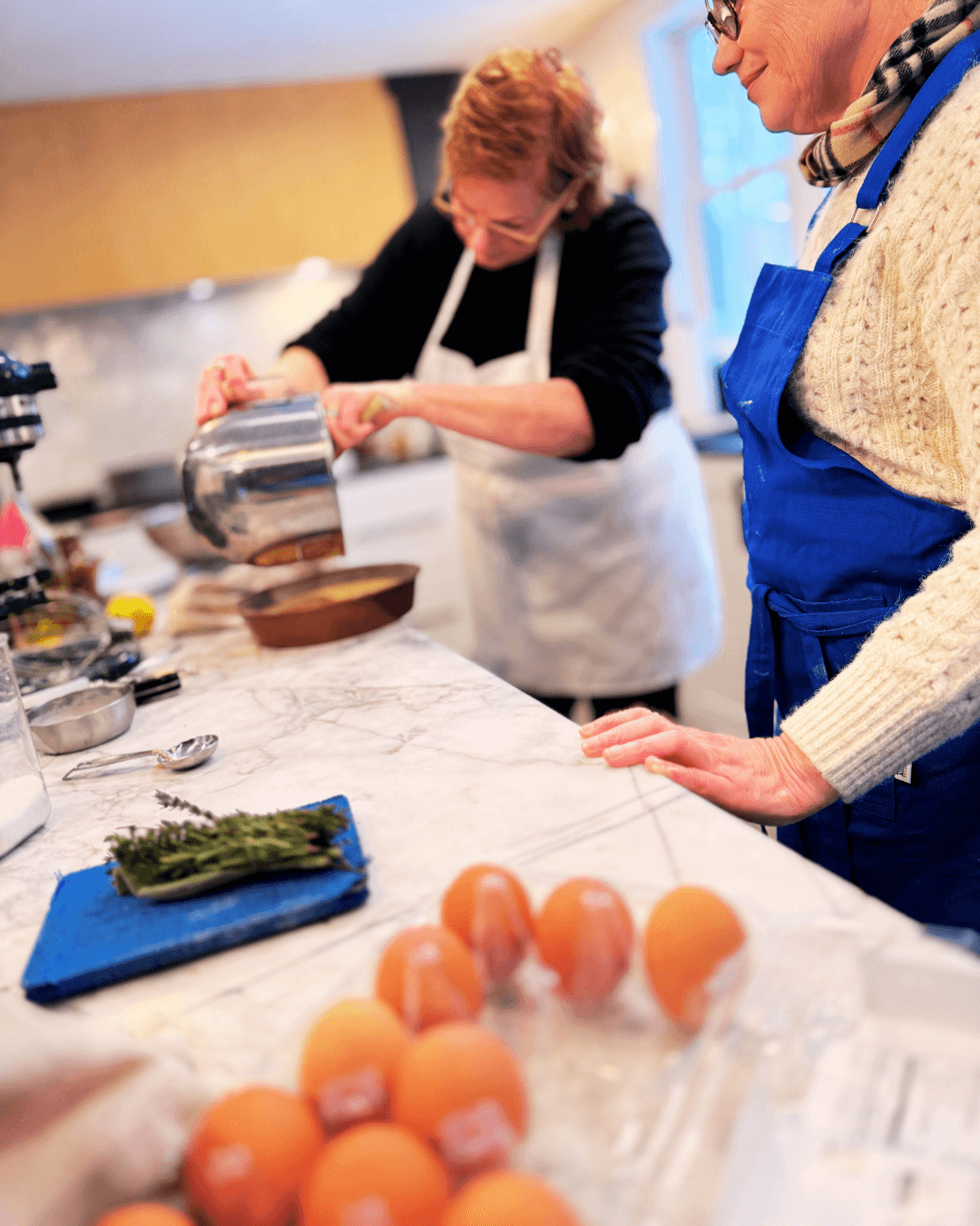 Two women cooking in a kitchen, with eggs and herbs on the counter, and one woman pouring ingredients into a bowl.