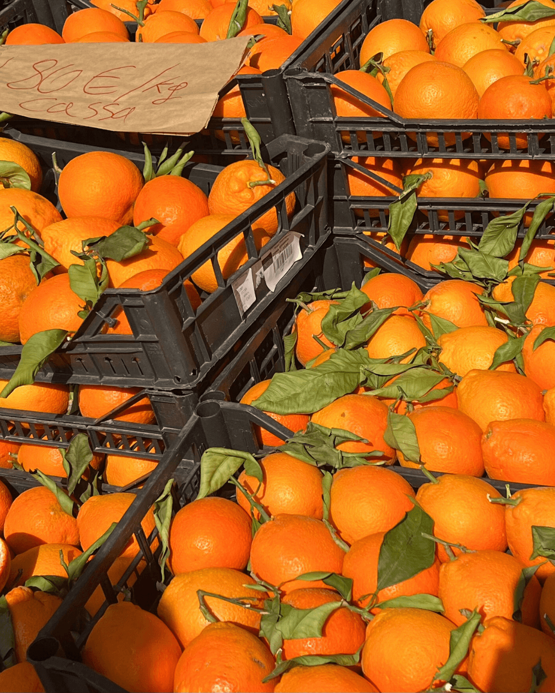 Crates filled with fresh oranges, some with green leaves attached, at a produce market.