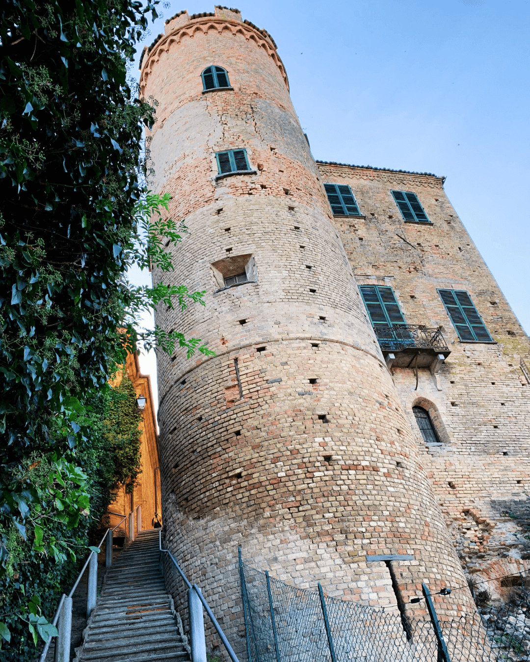 A tall, old brick tower with a circular structure, small windows, and a couple of blue shutters next to a building with similar brick walls and multiple blue-shuttered windows, taken from a low angle during the day.