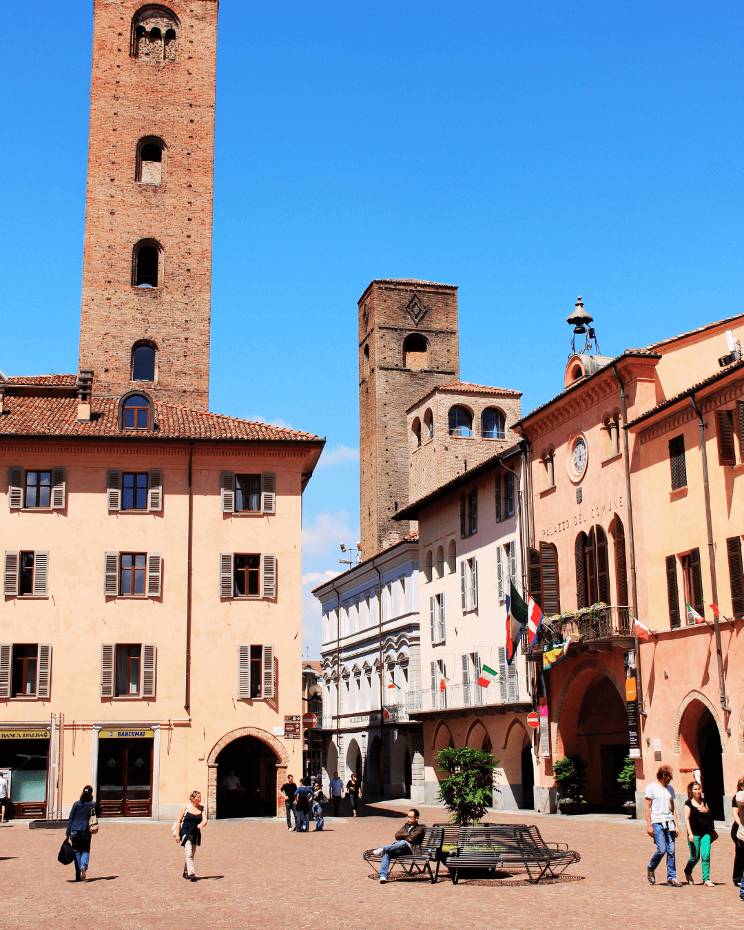 A lively European city square with historic brick towers, colorful buildings, flags, and people walking and sitting on benches under a clear blue sky.