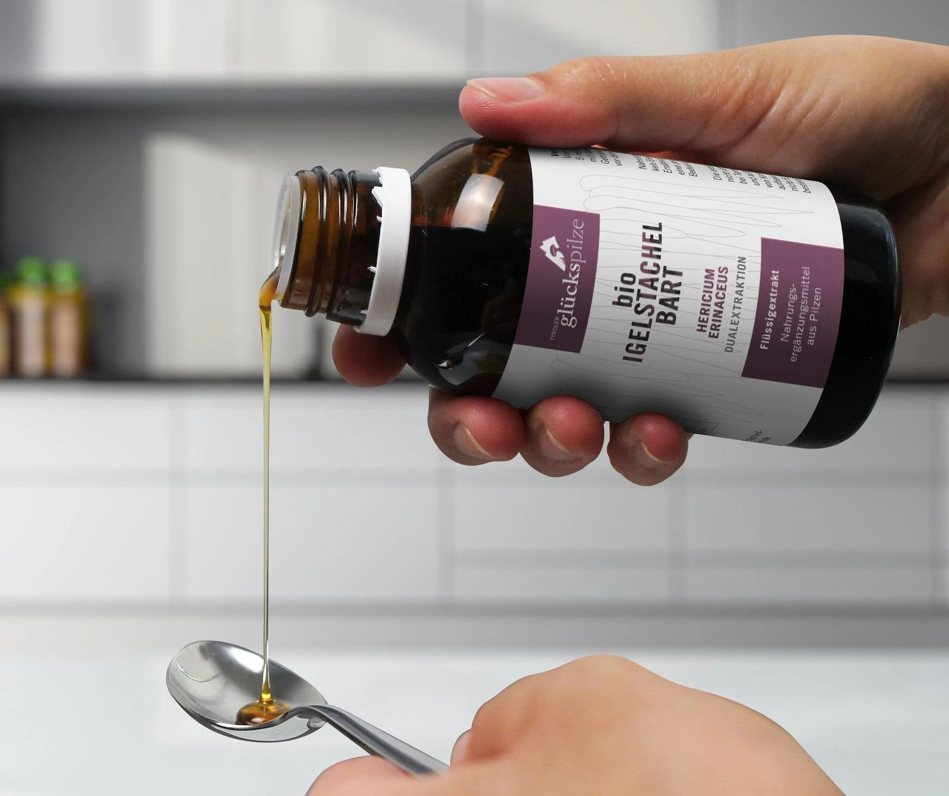Eine Hand hält eine braune Flasche mit Nahrungsergänzungsmittel, das Öl tropft in einen Löffel, im Hintergrund eine weiße Küchenwand mit einem Regal.