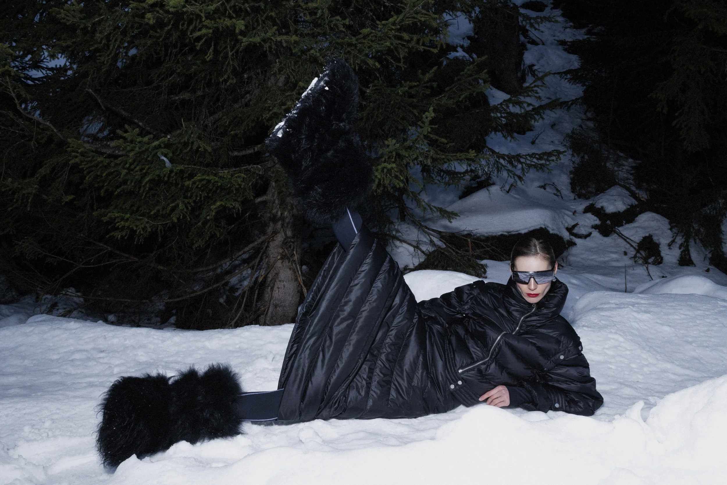 A woman wearing black sunglasses and a black jacket is lying on snow in a forested area with evergreen trees. She is resting on her stomach with her left arm bent under her head. There is snow on the ground and tree branches, and part of a person's black boot is visible in the upper left corner of the image.