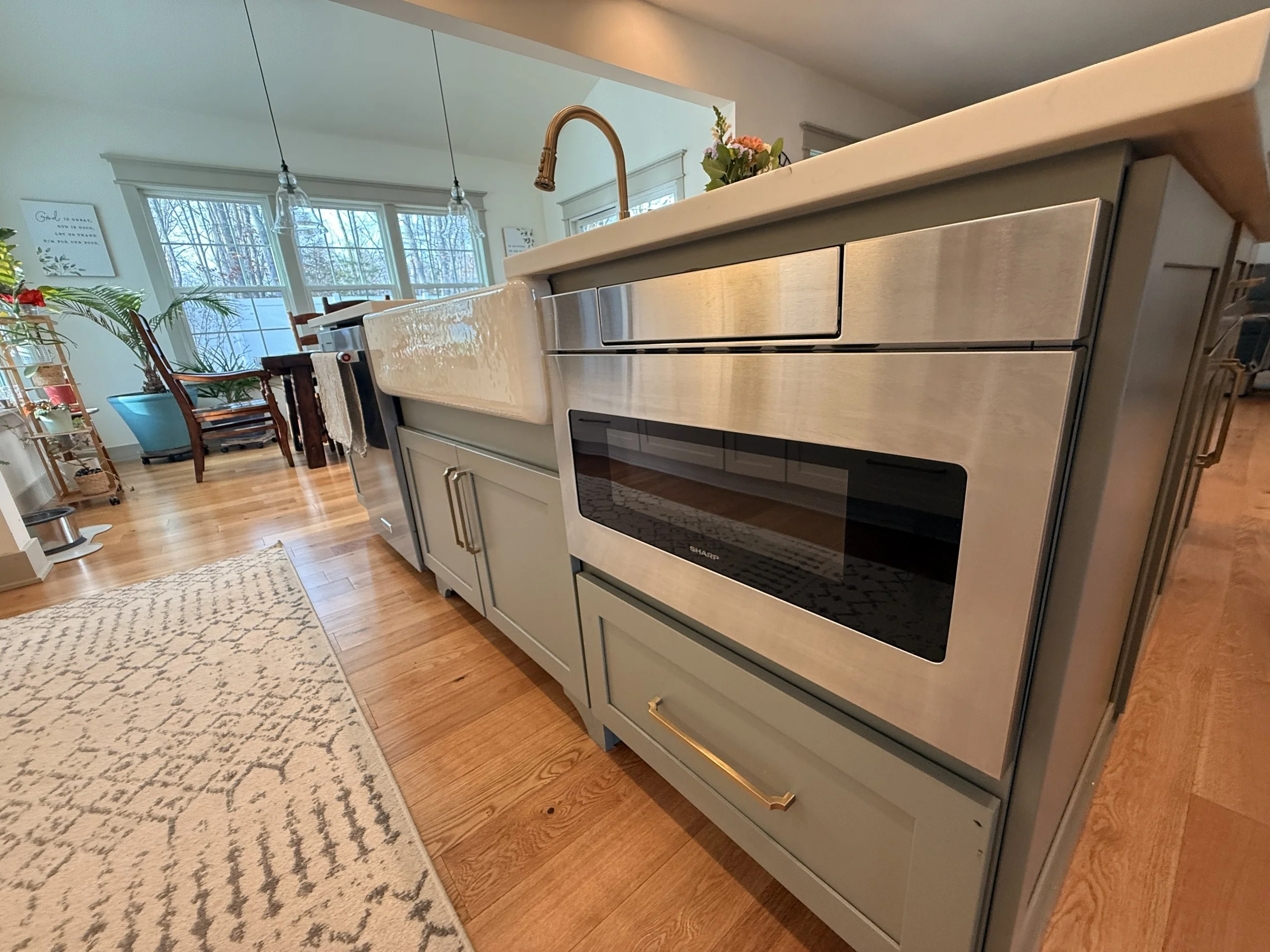 A modern kitchen with a light gray island, a stainless steel oven, a copper sink faucet, and a floral centerpiece. In the background, there is a dining area with large windows, wooden dining chairs, and hanging pendant lights, along with decorative wall art and indoor plants.