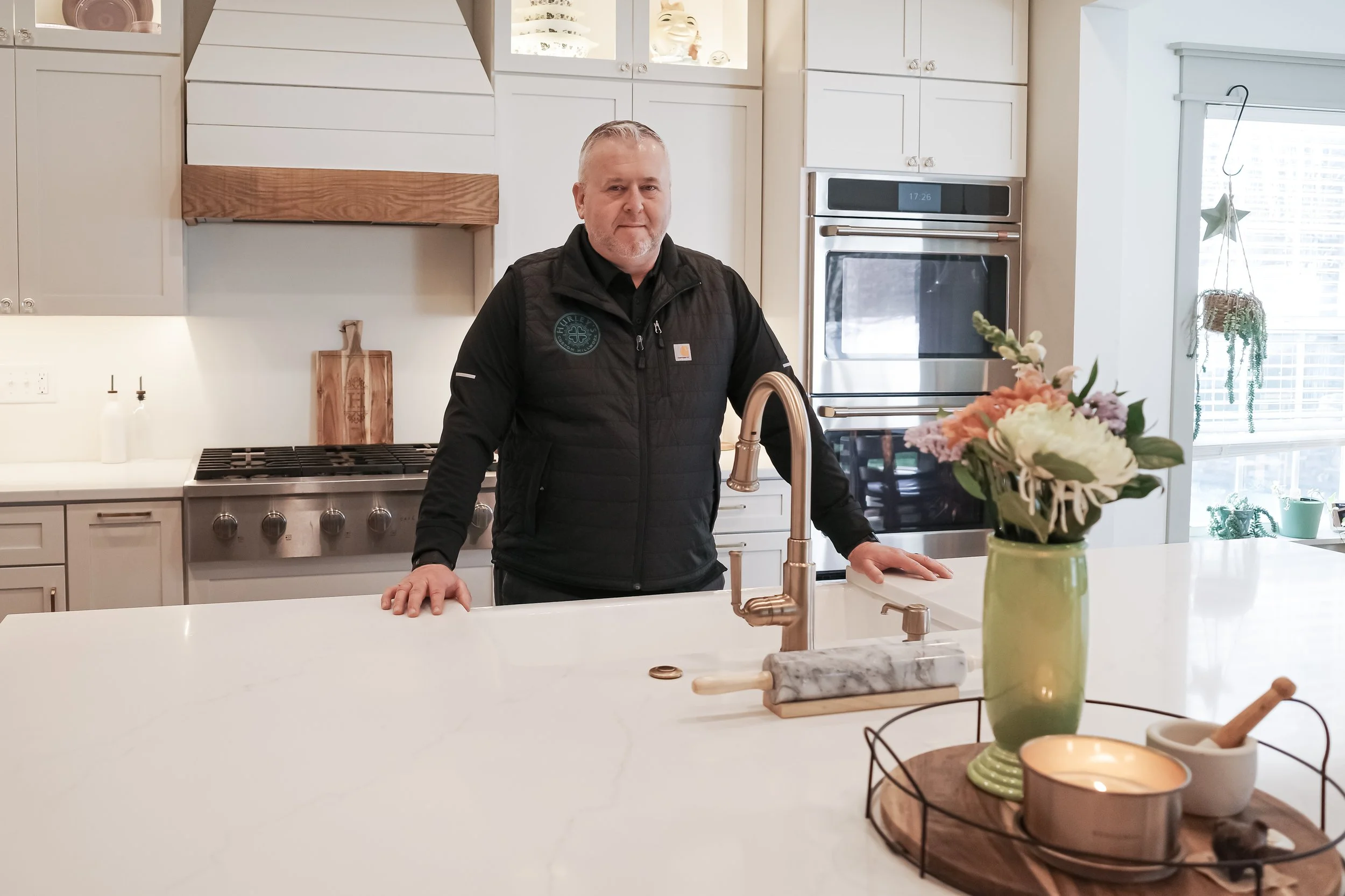 A man with gray hair and a beard wearing a black vest stands in a kitchen next to a large white island with a potted flower in the foreground. The kitchen has white cabinets, a stainless steel oven, and a large window with a star-shaped decoration hanging from the blinds.