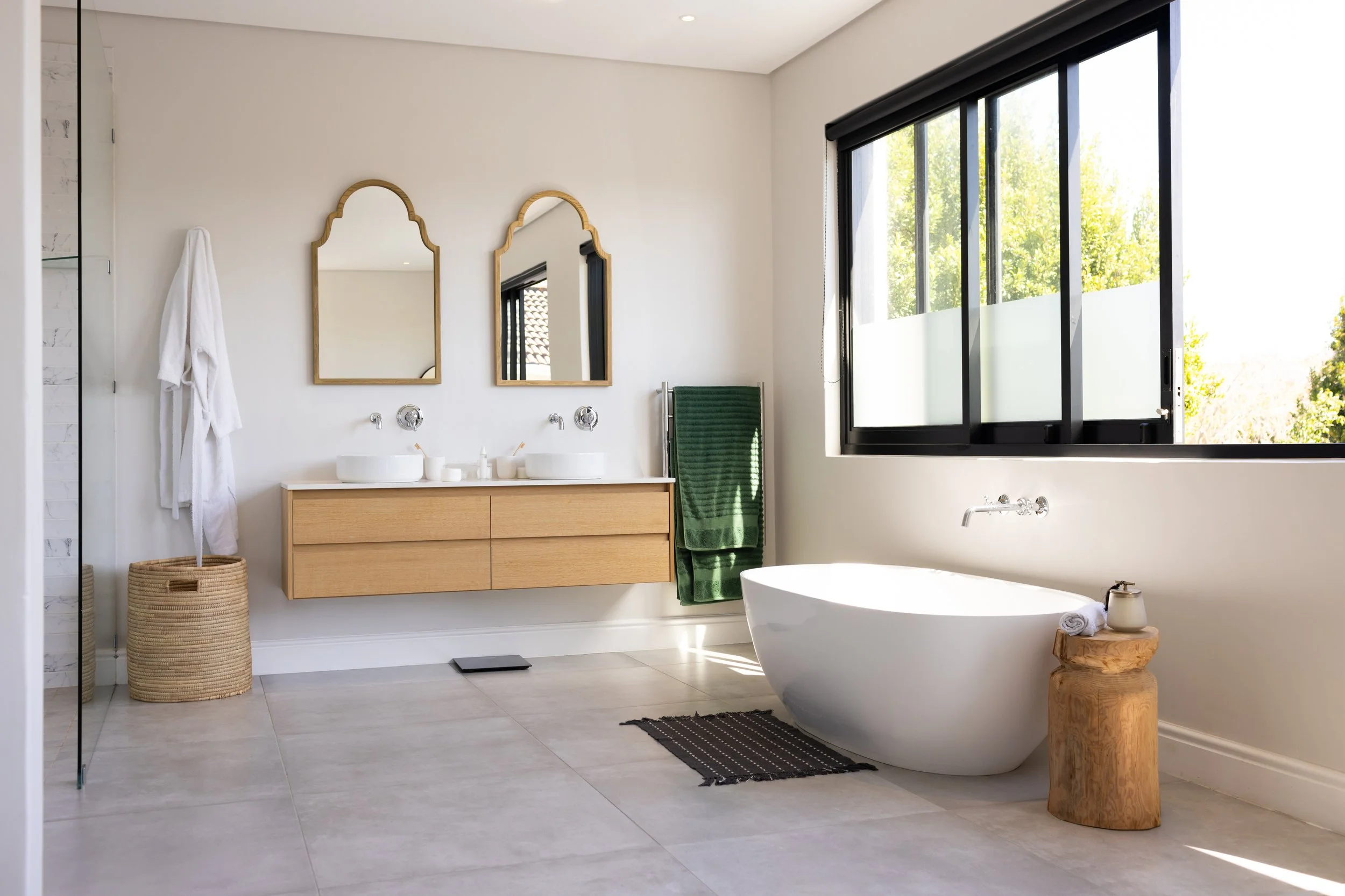 Modern bathroom with a freestanding bathtub, two sinks with mirrors, a window with black frame, and minimal decor.