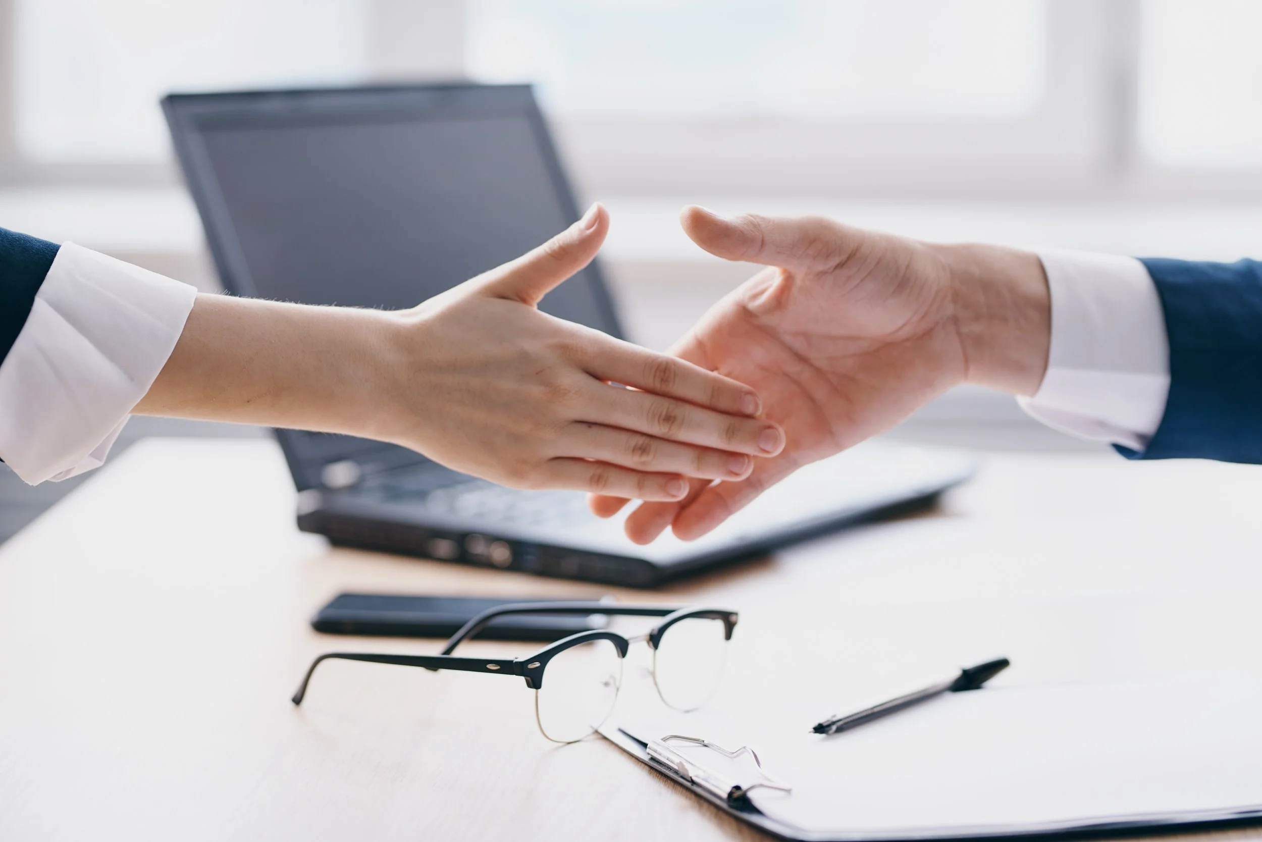 Two people shaking hands over a desk with a laptop, glasses, pen, and papers.