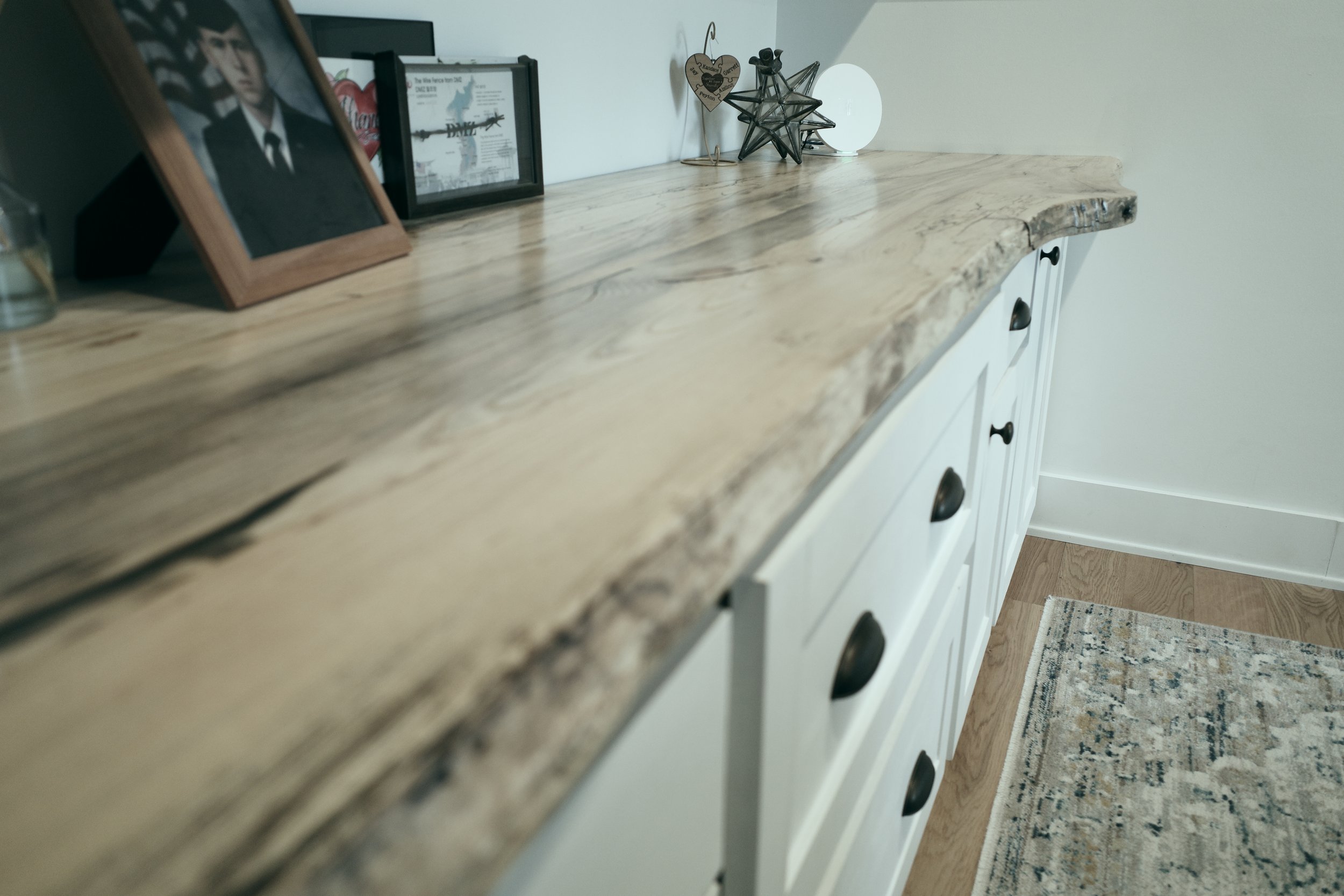 A kitchen or dining area with a wooden countertop, white cabinets with black handles, a patterned rug, and decorative items including framed photos, a heart-shaped wooden decor piece, a star-shaped decor made of metal, and a white round object on the countertop.