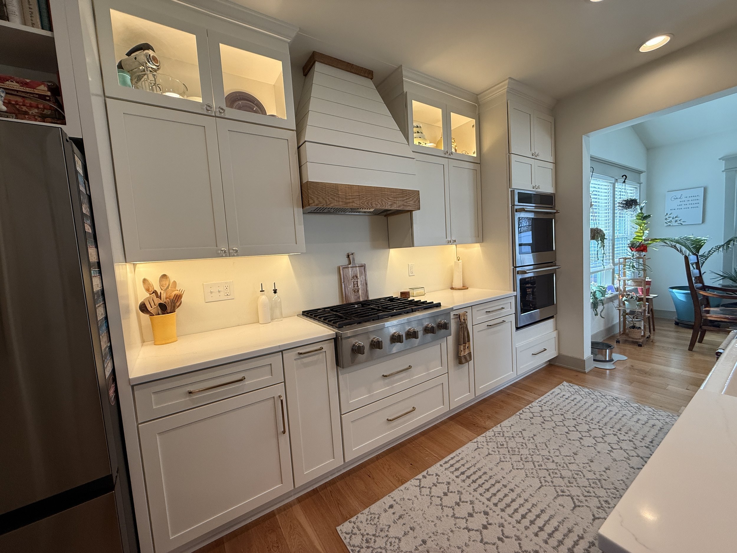 A kitchen with white cabinets, a gas stove, and a wooden range hood, located next to a dining area with a large window and plants.