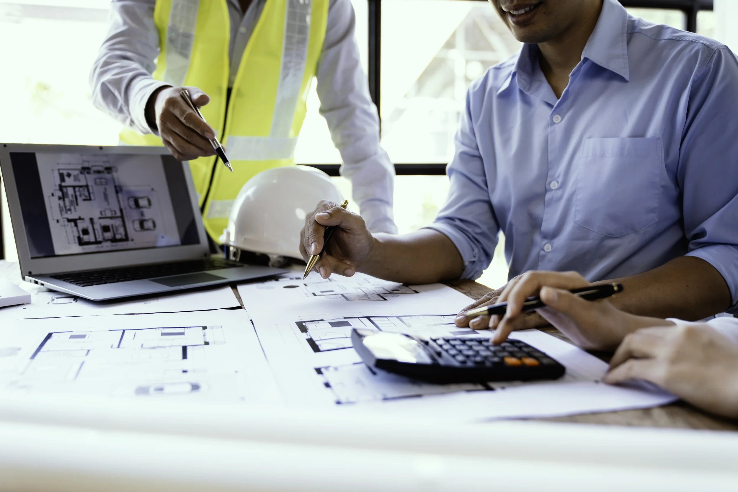 Two engineers working on architectural plans at a table with a laptop, a white safety helmet, a calculator, and a smartphone, in a well-lit office with large windows.