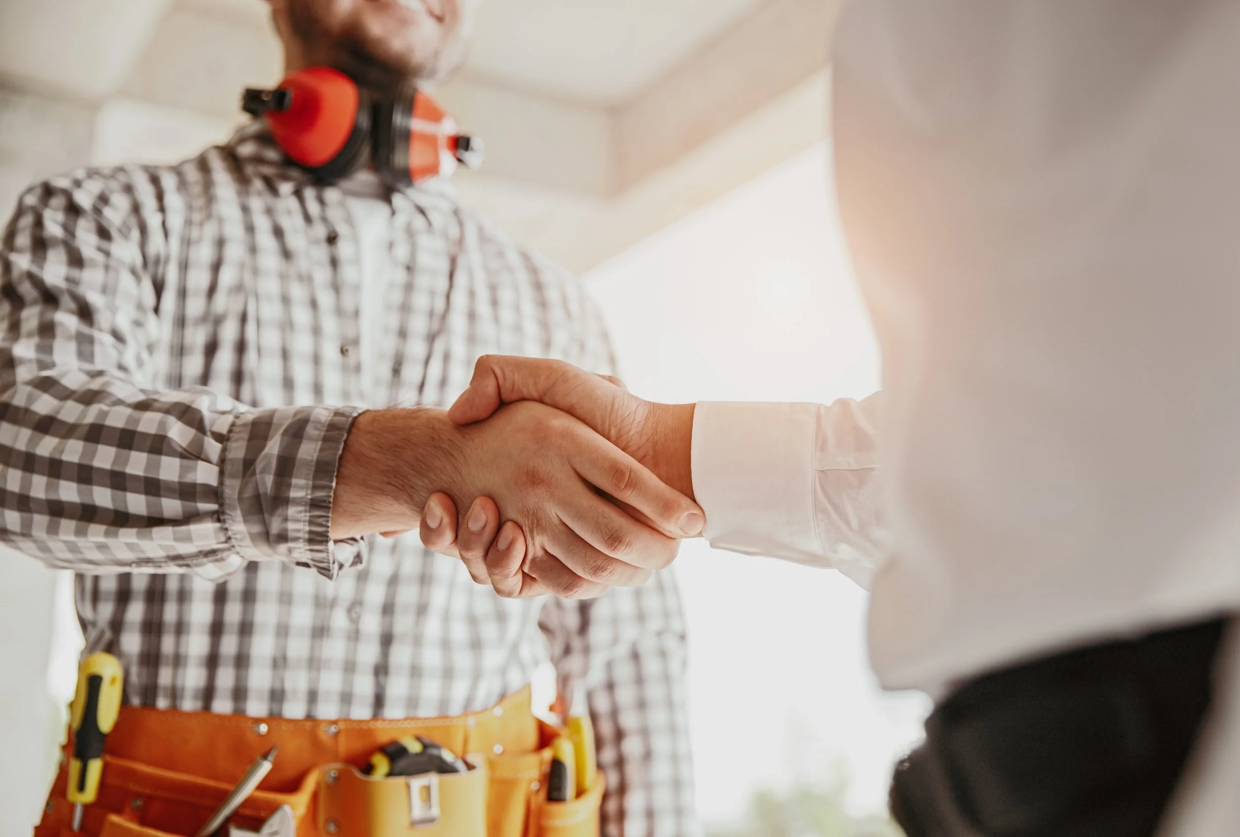 A handyman wearing a plaid shirt and a tool belt shaking hands with a person in a white shirt inside a building.