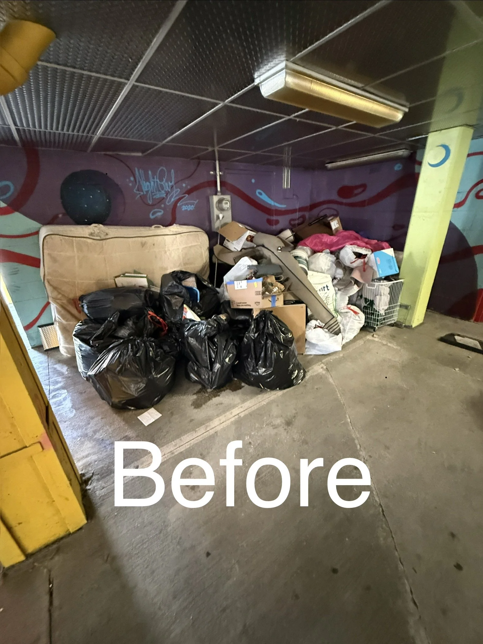 A cluttered underground space with trash bags, cardboard boxes, and miscellaneous items on the floor. There is graffiti on the purple wall, and fluorescent lights on the ceiling. The floor is concrete.
