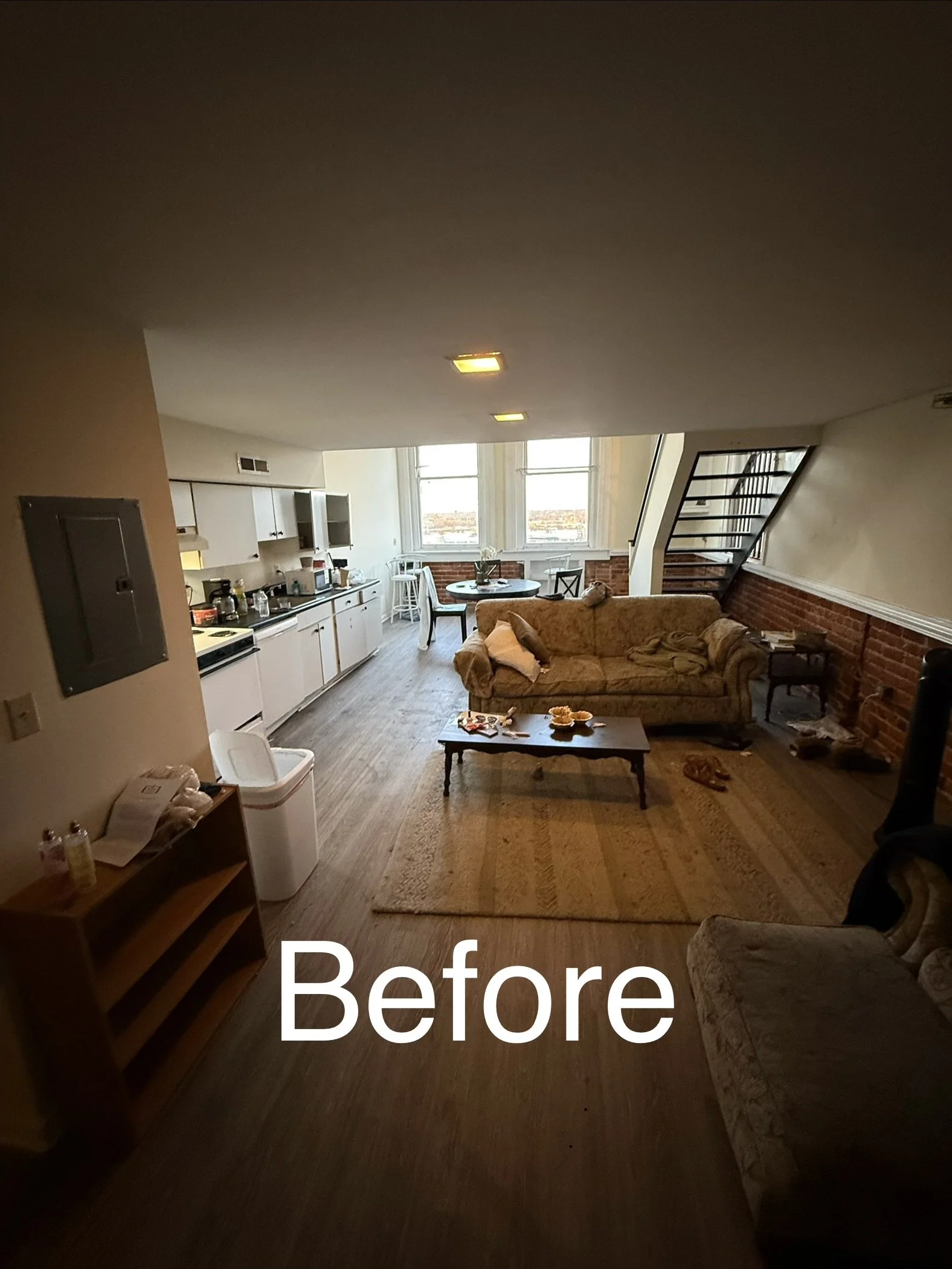 Living room with a sofa, coffee table, and kitchenette near a large window, with stairs on the right side.