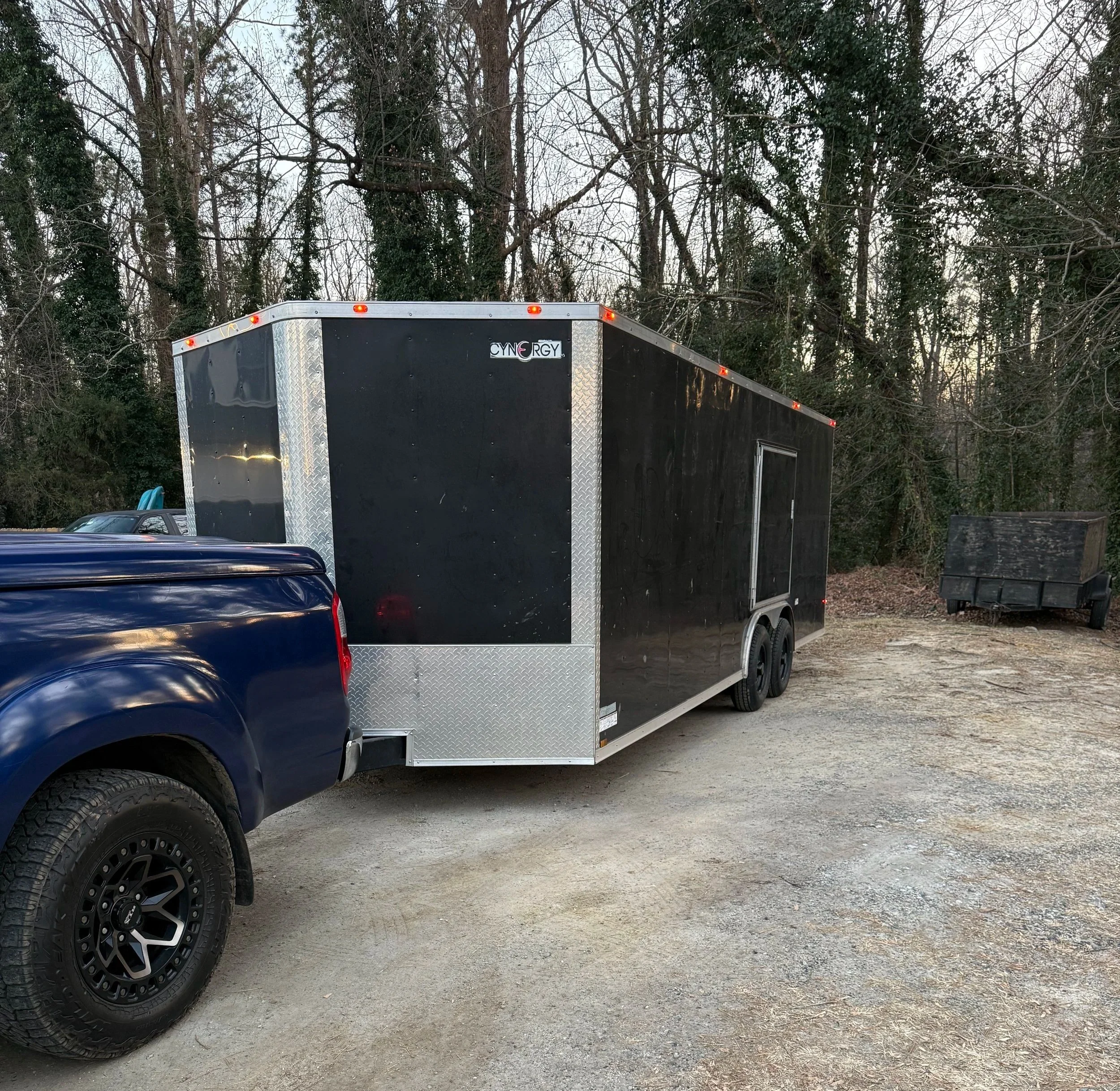 Black enclosed trailer with a sticker that says "CYNGRY" on the upper part, parked outdoors against a backdrop of leafless trees.