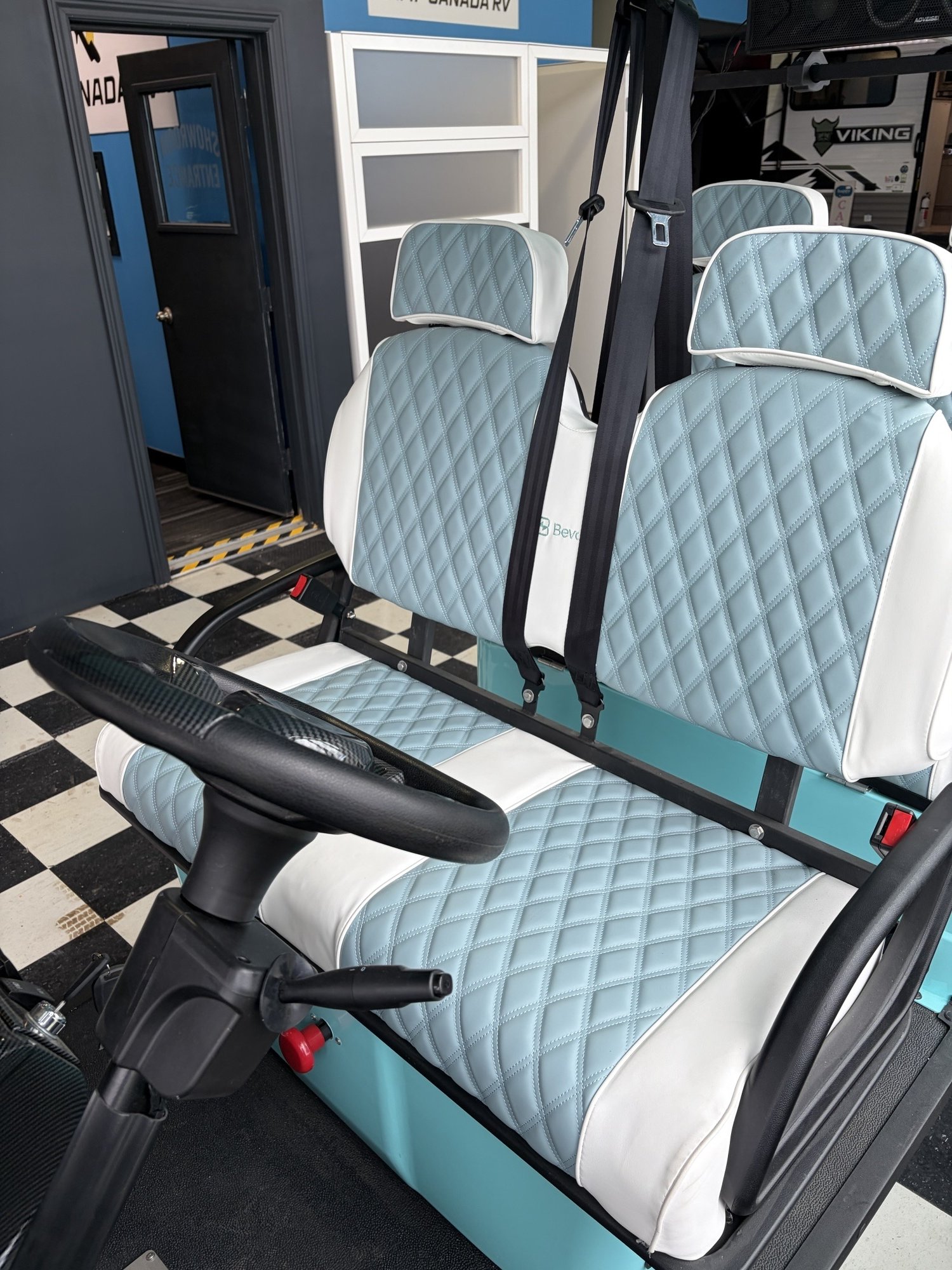 A golf cart with a light blue and white diamond-stitched seat and backrest, located indoors with checkered flooring, in front of a black door and a white partition.
