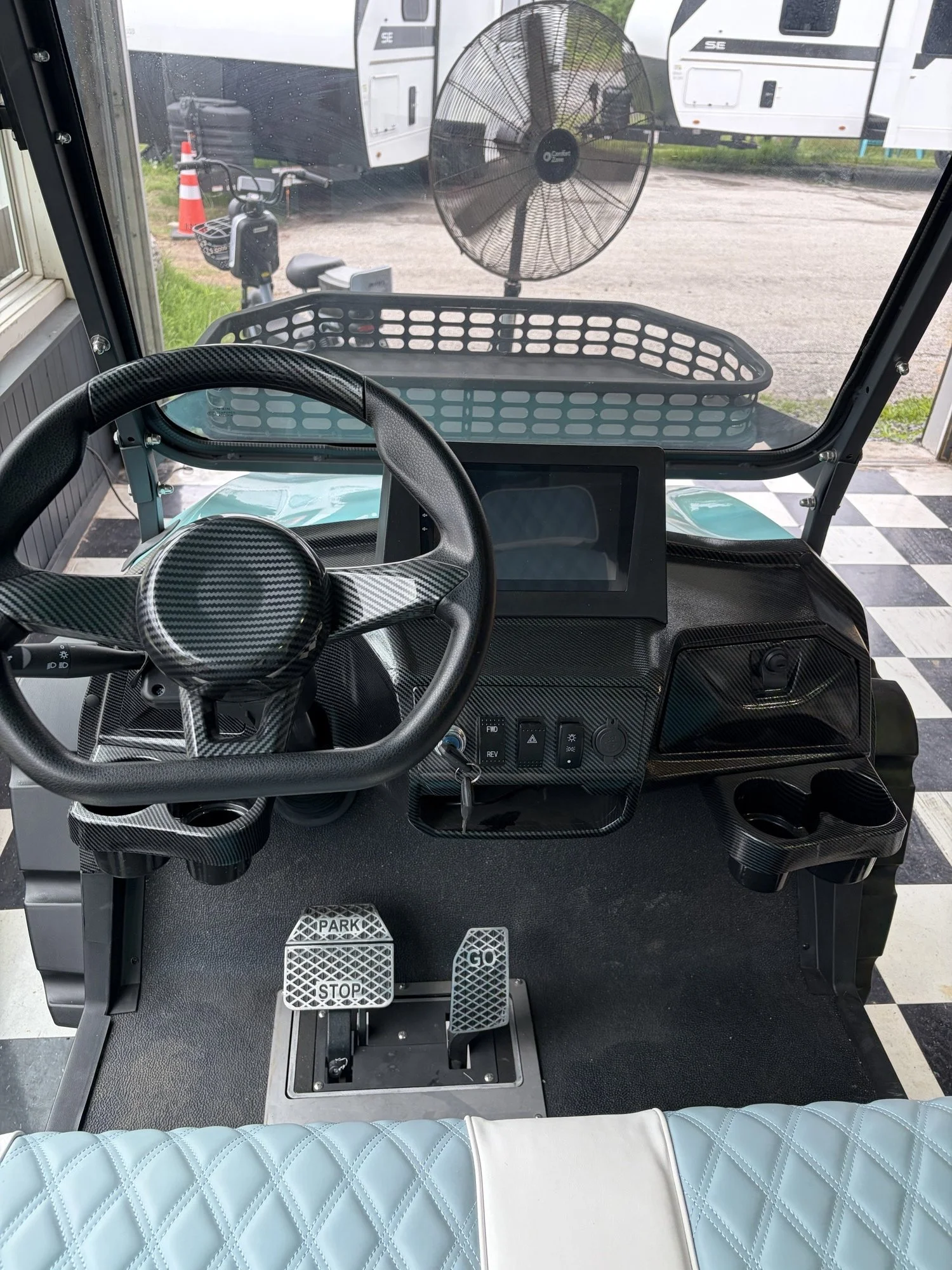 Interior of a golf cart showing the steering wheel, dashboard with some controls, and pedals labeled 'STOP', 'GO', and 'SPEED'. Outside, a baseball field is visible.