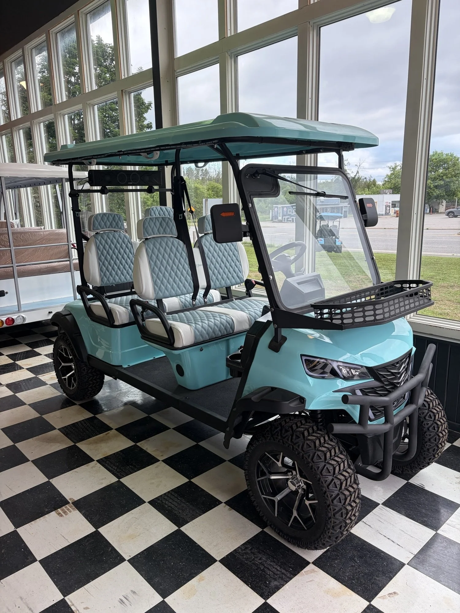 A turquoise golf cart with four cushioned seats, parked indoors on black and white checkered flooring next to large windows showing an outdoor view of trees, parked cars, and a cloudy sky.