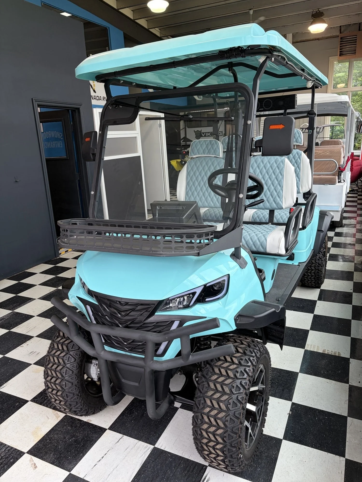 A blue golf cart with a clear windshield and quilted upholstery seats inside a showroom with black and white checkered flooring.