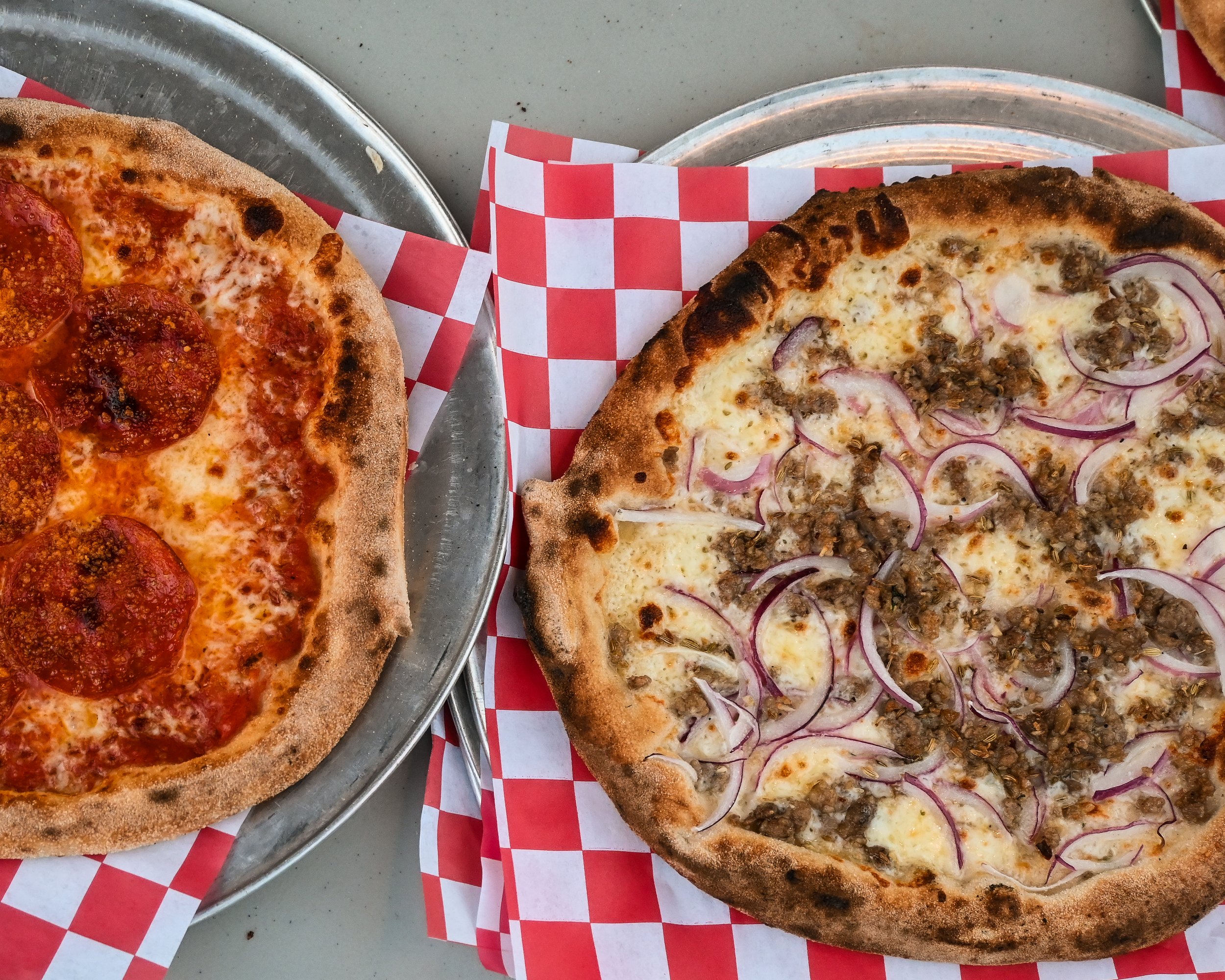 Two pizzas on metal trays with red and white checkered paper at an outdoor catering event.