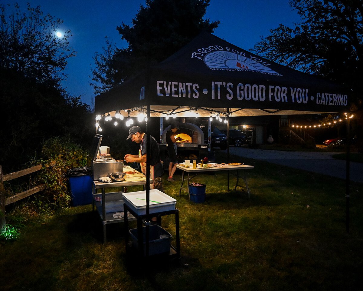 Nighttime outdoor catering setup with a black tent, string lights, and Alessandro preparing food on tables with a pizza oven in the background.