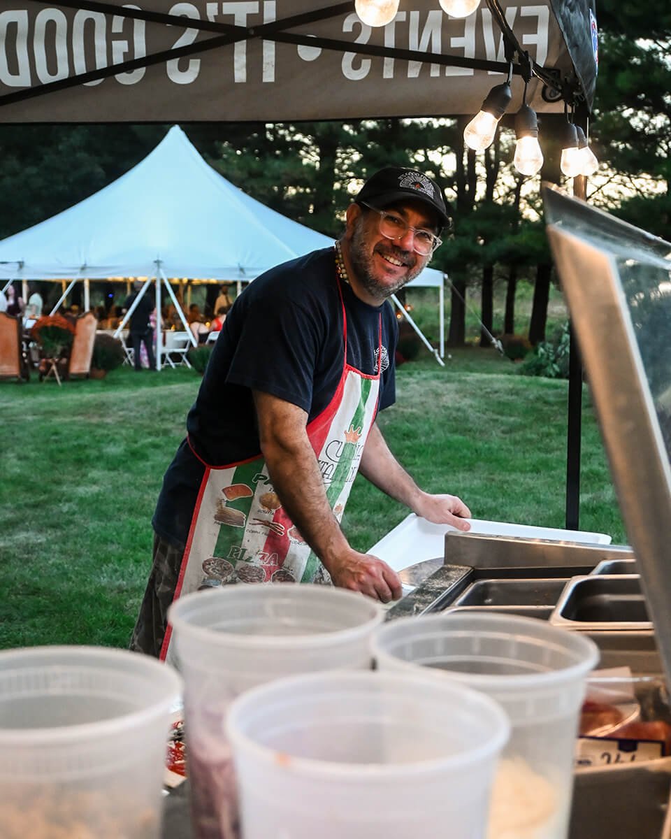 Outdoor catering setup with a black tent, string lights, and Alessandro preparing food on tables with a party tent in the background.