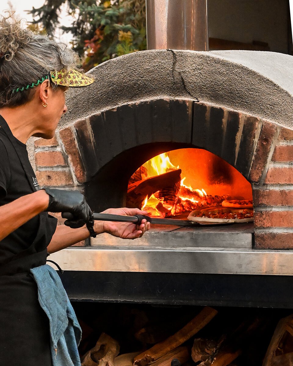 A woman placing a pizza into a wood-fired brick oven with a roaring fire inside at an outdoor catering event.