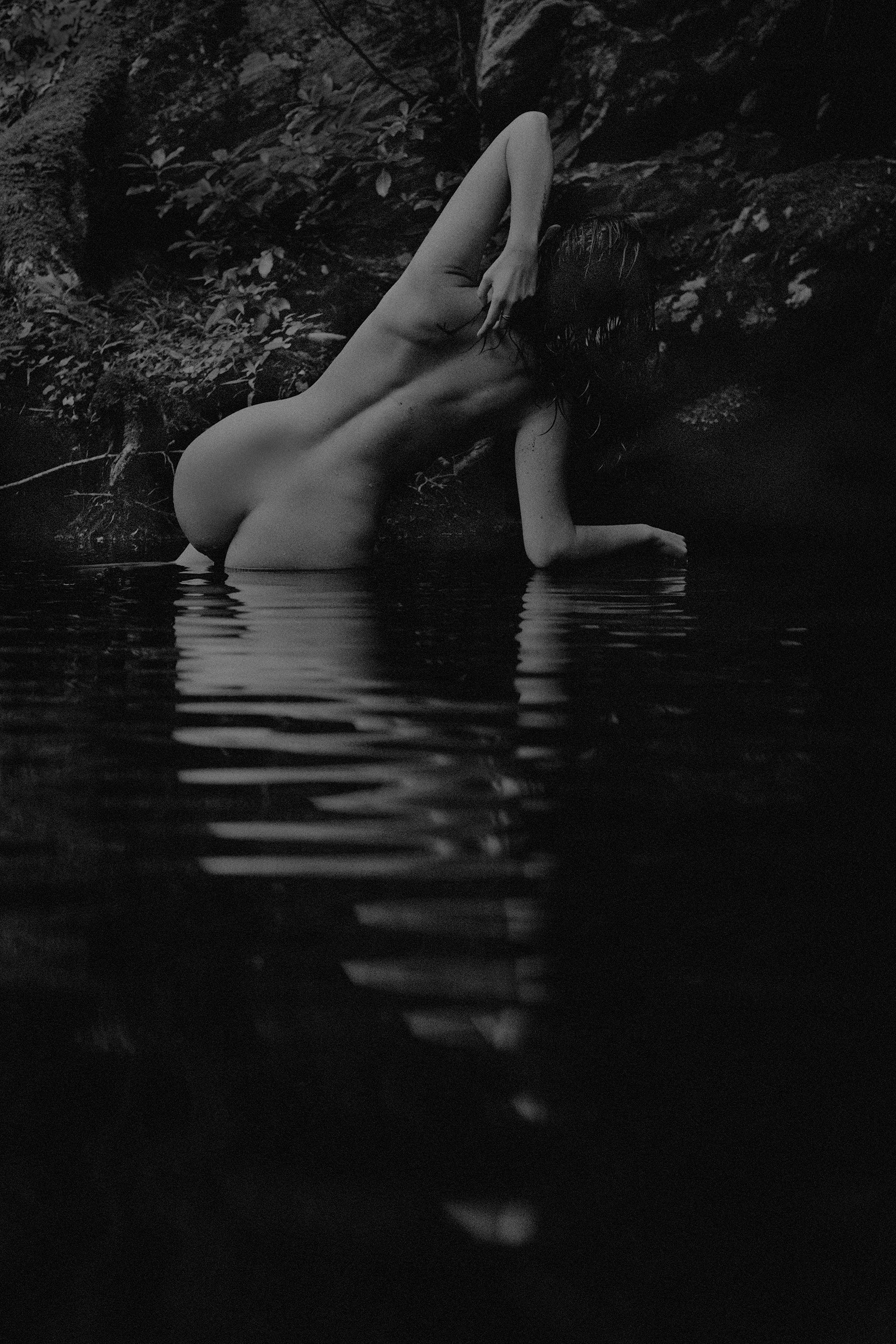 A black and white photograph of a nude woman with wet hair, partially submerged in water, leaning against a rock or tree in a forested area.