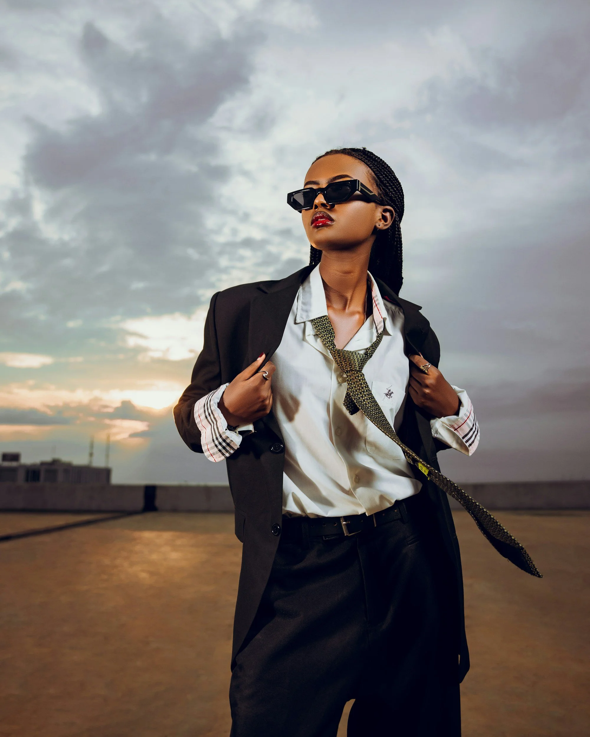 A woman wearing a black blazer, white shirt, and black pants stands outdoors during sunset, with dark cloudy skies overhead. She has braided hair, sunglasses, and a loosely tied necktie, exuding a confident demeanor.