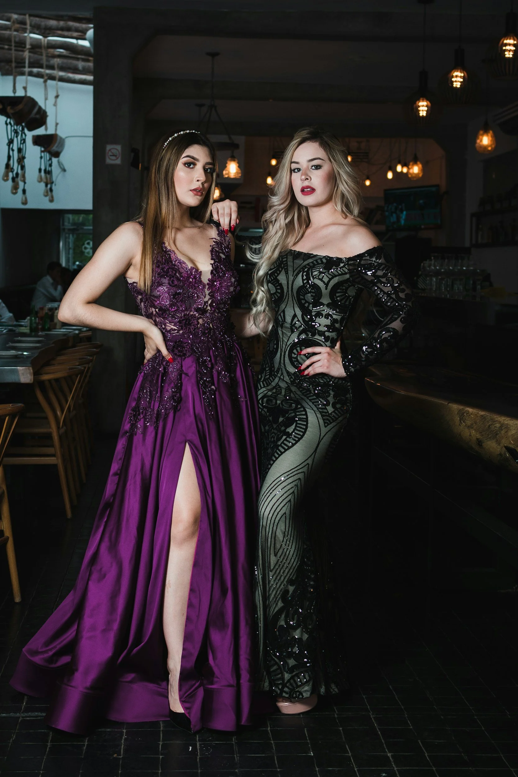 Two women in elegant evening dresses standing inside a restaurant or bar, posing for a photo. The woman on the left wears a purple dress with lace details and a high slit, while the woman on the right wears a black and green patterned dress with sequins.