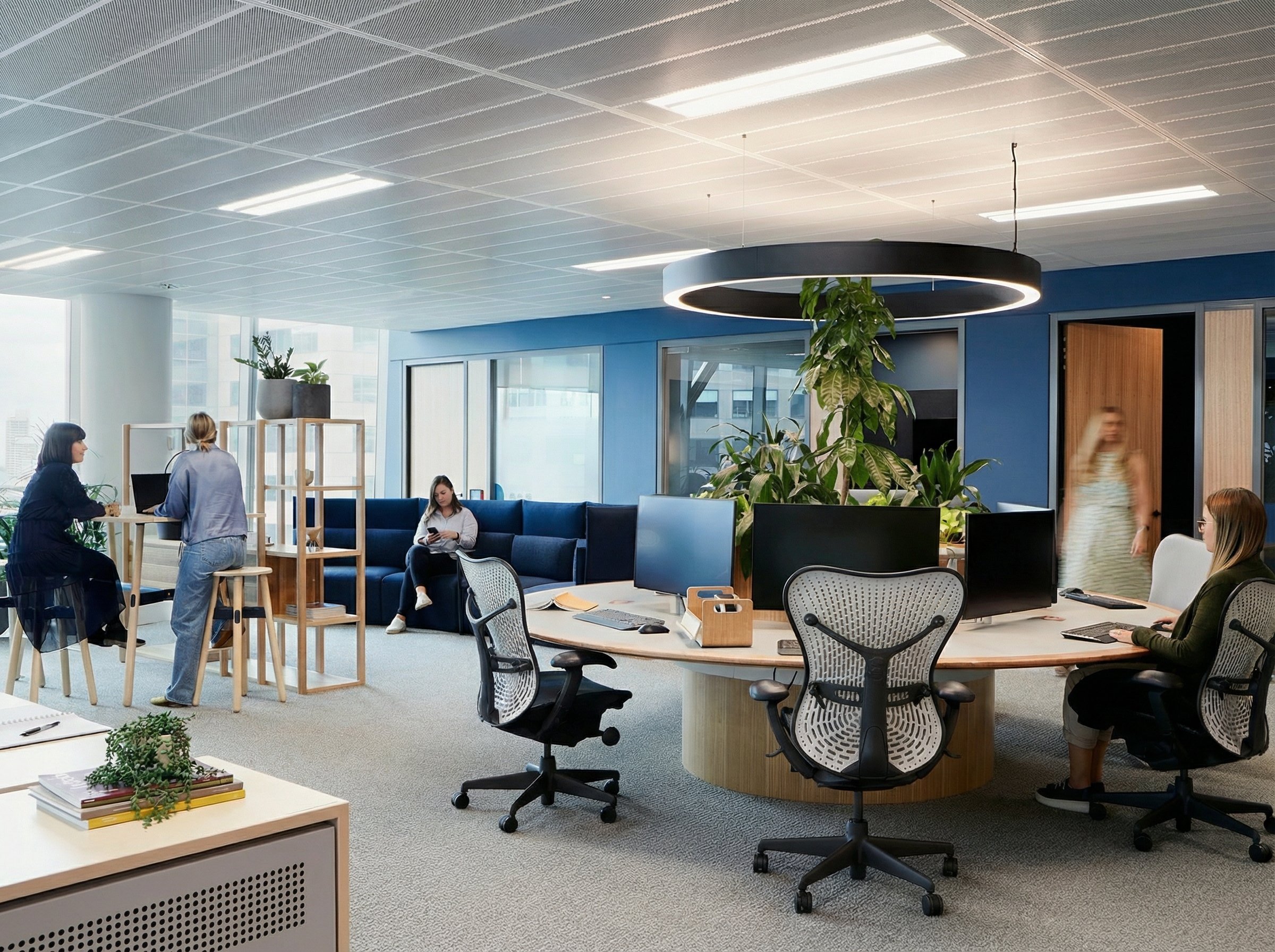 Modernes Büro mit mehreren Mitarbeitenden, die an Computern arbeiten, sich unterhalten oder Pausen machen, mit großen Fenstern, blauen Wänden und vielen Pflanzen.