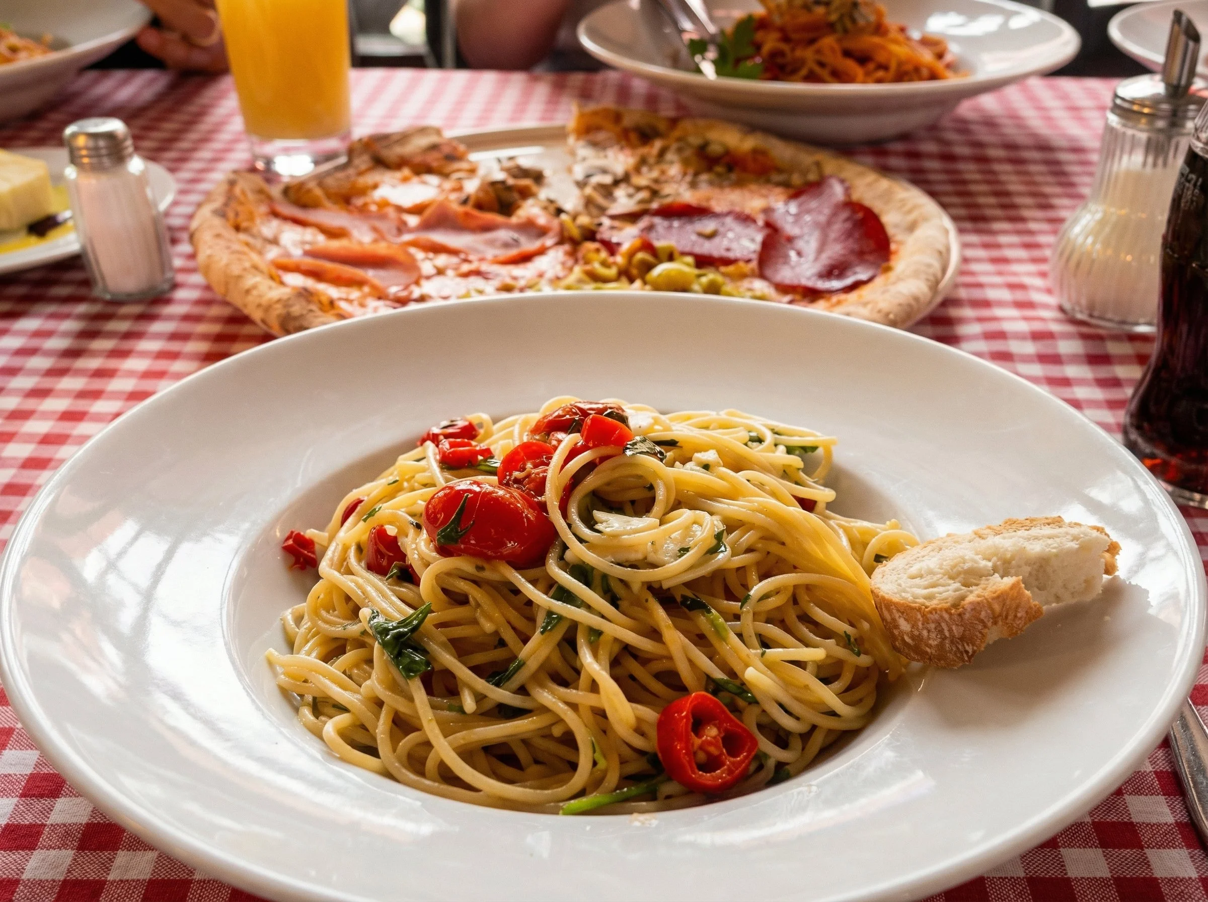 Pasta mit Kirschtomaten, Basilikum und Chili auf einem weißen Teller, im Hintergrund Pizza und Salat auf einem Tisch mit rotem und weißem Karomuster.