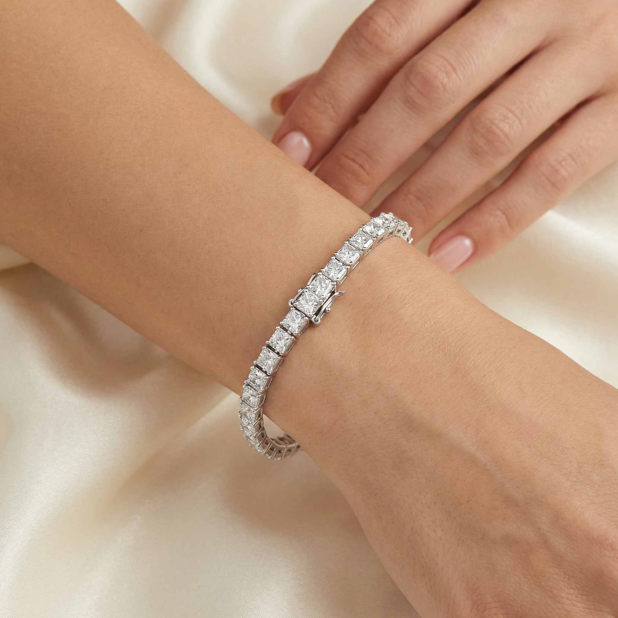 Eine Hand trägt ein schmales Armband mit großen, quadratischen, klaren Steinen, auf einem hellen Satin-Hintergrund.
