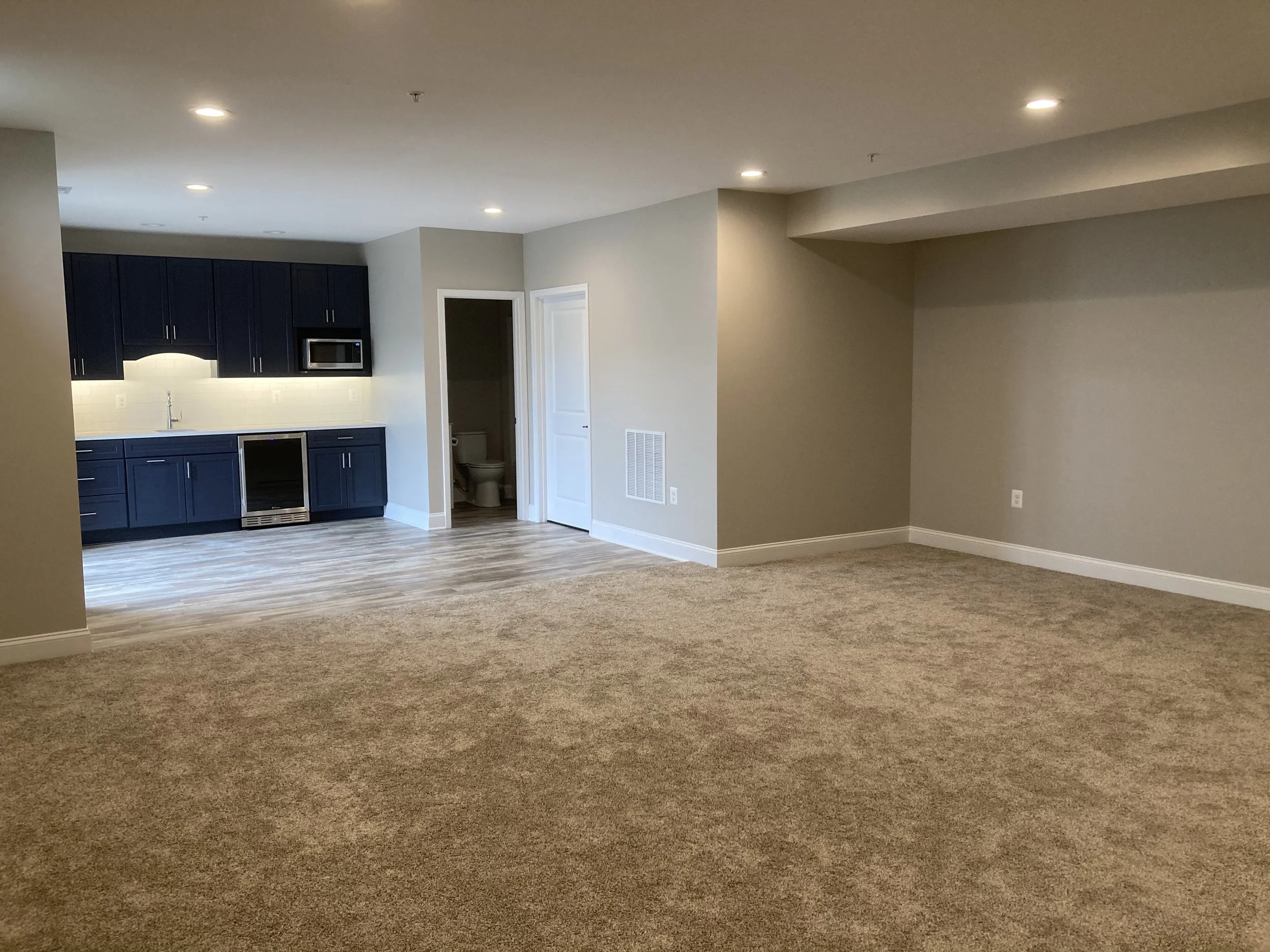 Finished basement space with beige carpet, gray walls, and ceiling lights, with kitchen area featuring blue cabinets, a stainless steel microwave, and bathroom.