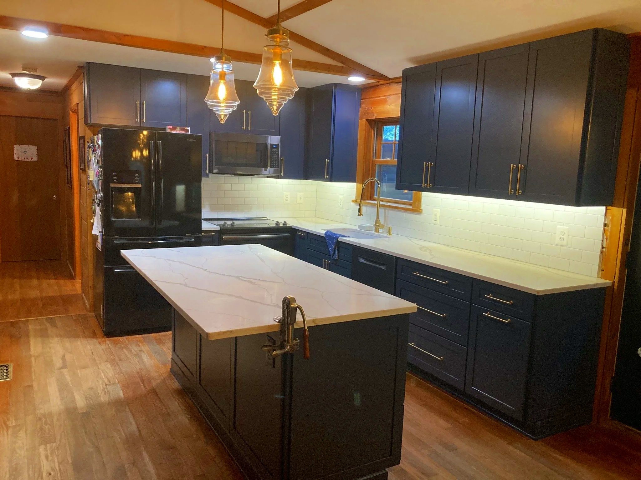 Modern kitchen with black cabinets, white marble countertops, a black refrigerator, microwave, and a window above the sink with a gold faucet, wooden accents, and hardwood floors.