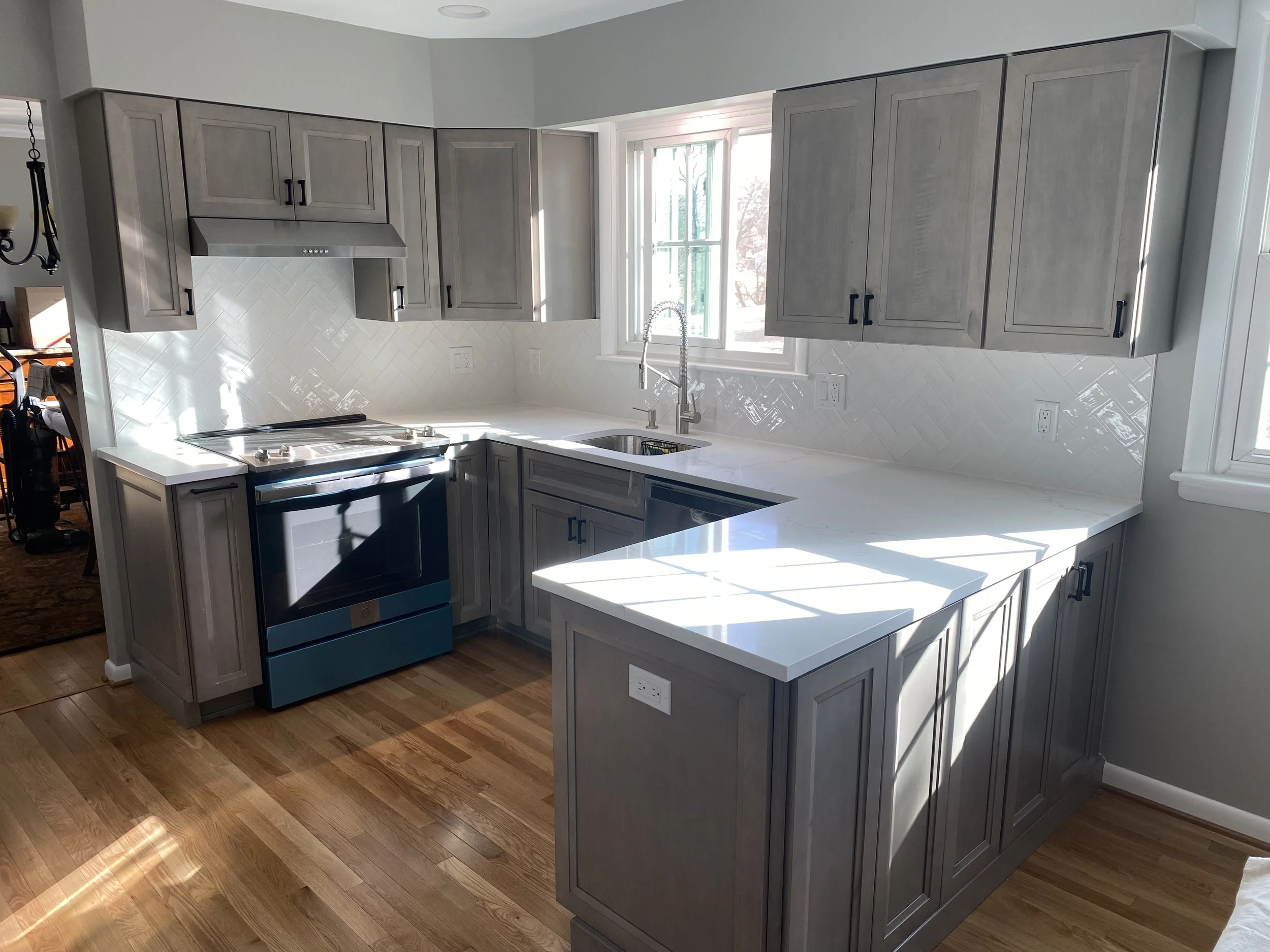 Bright kitchen with gray cabinets, white countertops, white tile backsplash, window above sink, and hardwood floor.