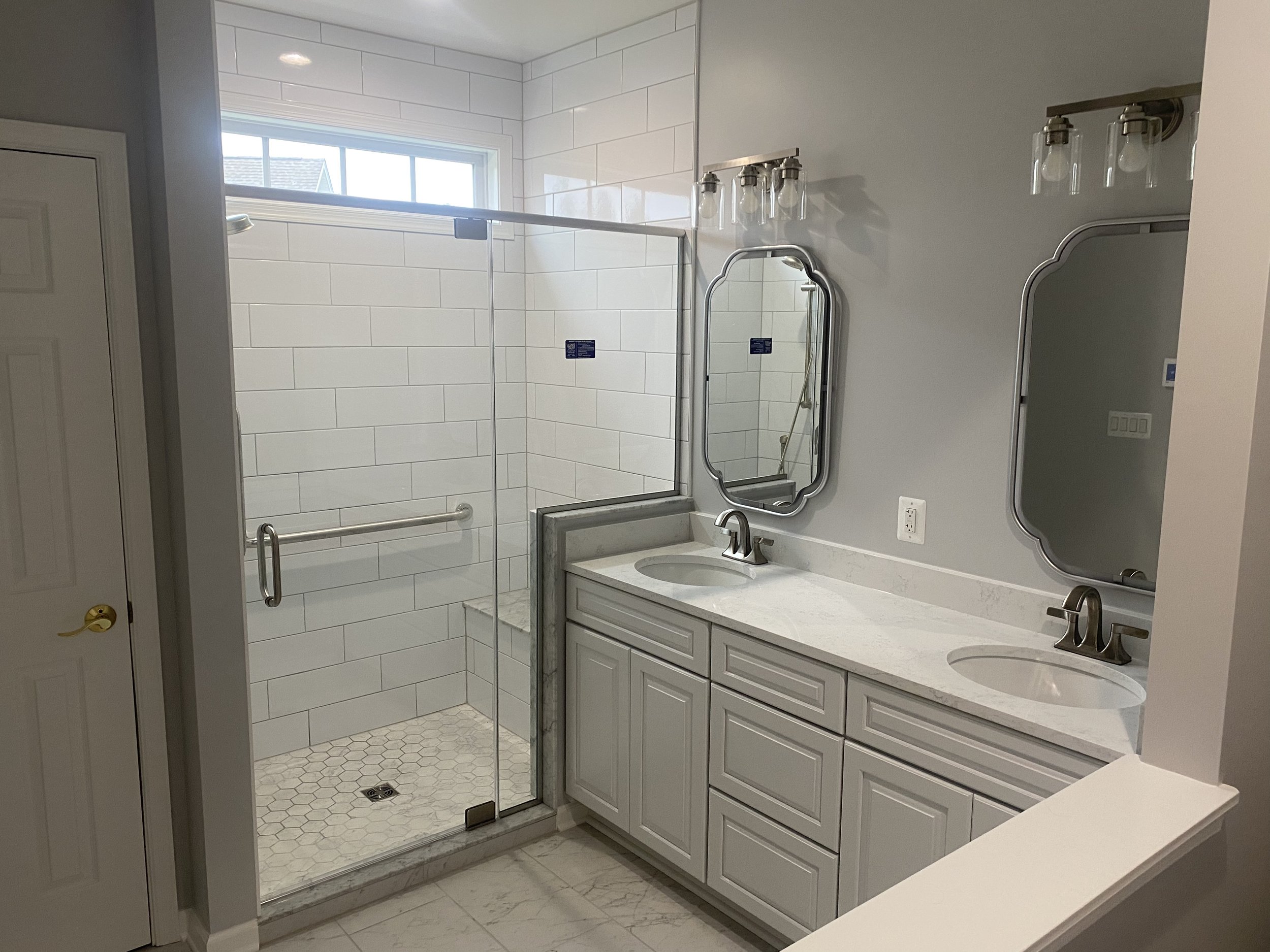 Bathroom with a double vanity featuring two mirrors and light fixtures, and a walk-in shower with a glass door and white subway tiles.