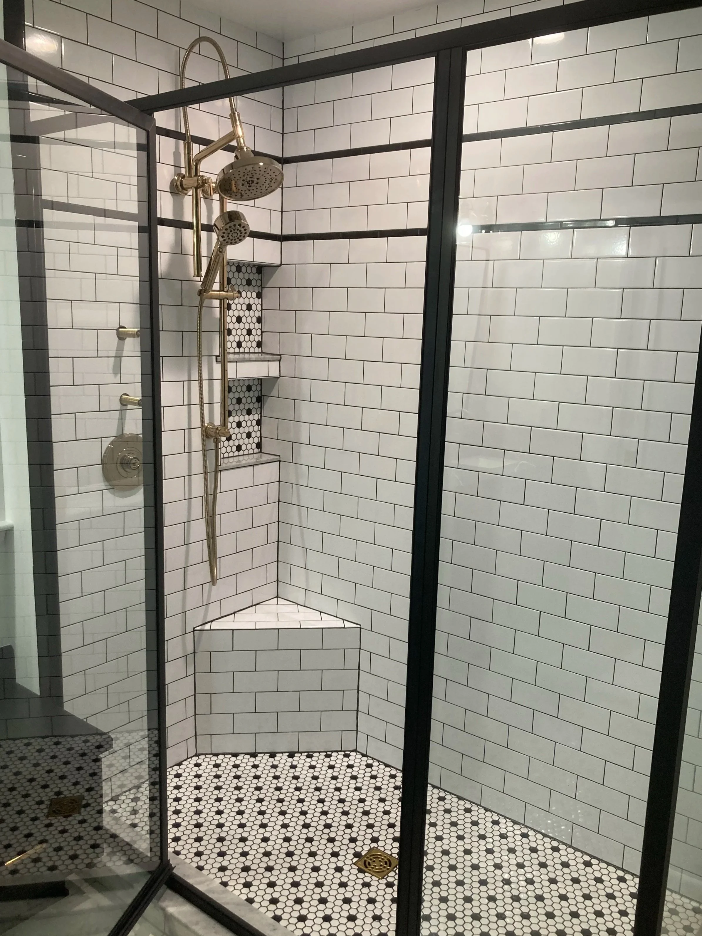 A modern walk-in shower with white subway tiles on the walls, black grout, and a black metal frame. The shower floor has small black and white hexagonal tiles, and there is a built-in white corner seat with a black mosaic tile accent behind it. The shower features a gold-colored showerhead, a hand shower, and several controls.