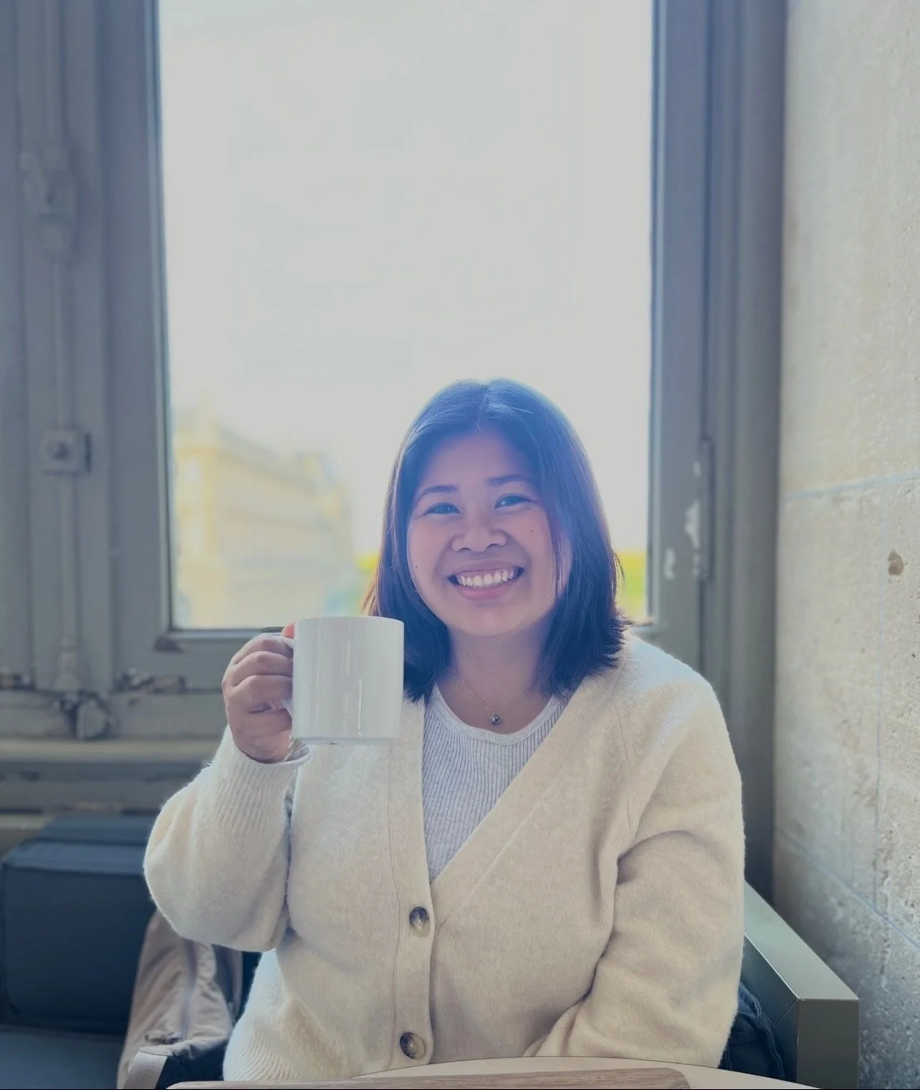 A smiling woman with shoulder-length dark hair holding a white mug, sitting in front of a window during daytime.