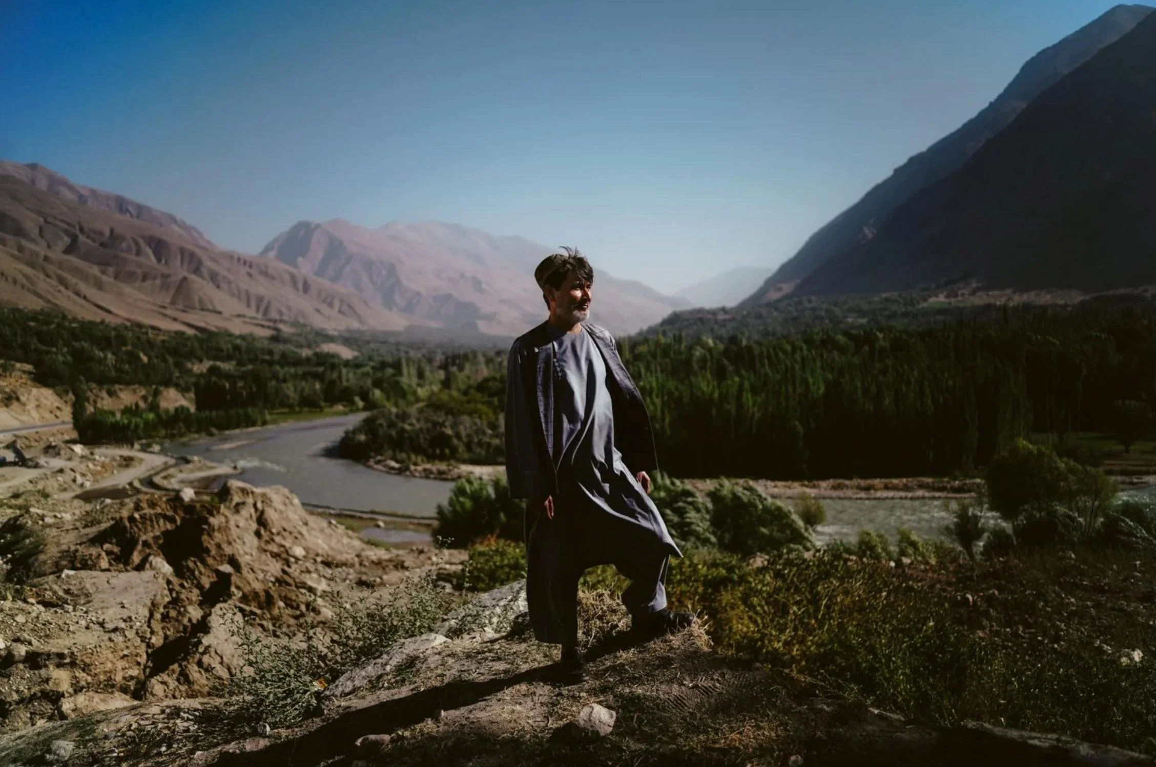 A man standing on rocky terrain overlooking a river and lush green valley surrounded by mountains under a clear blue sky.