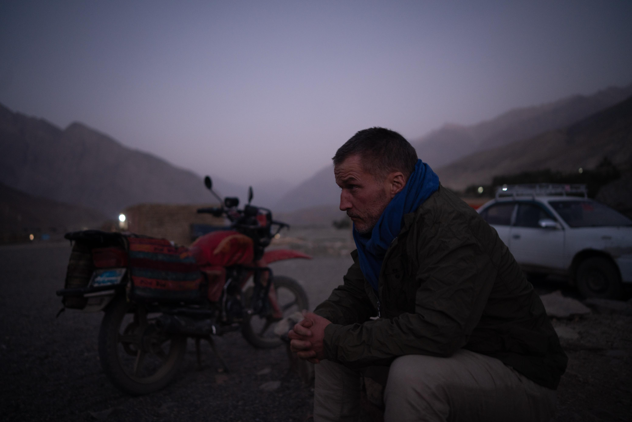 Ben Timberlake sitting outdoors with mountains in the background during dusk, next to a motorcycle and a car.
