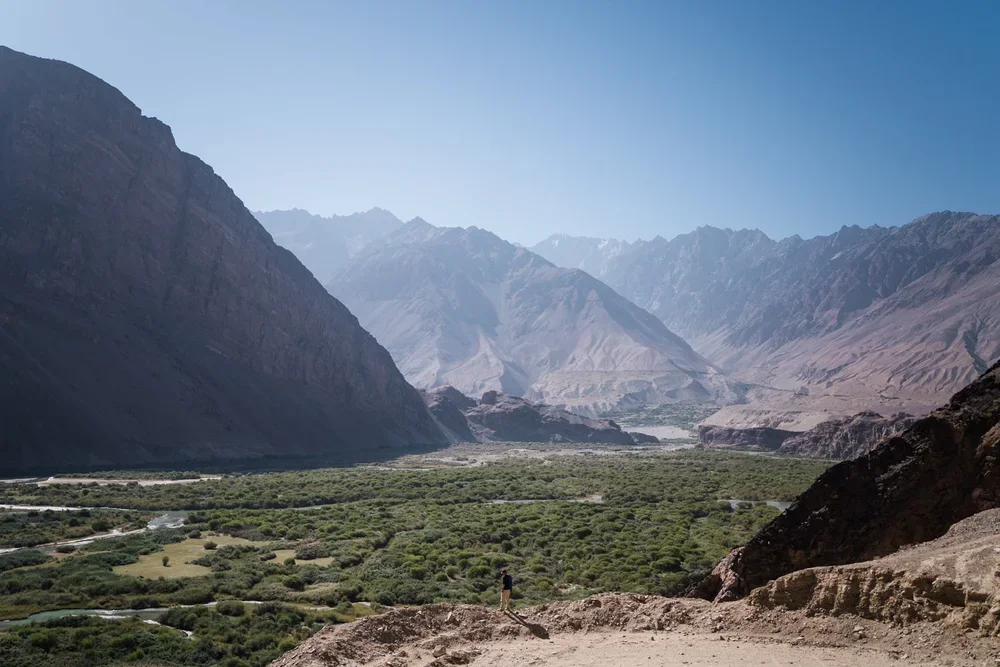 Scenic view of mountains, valley with greenery, and a person standing on a trail.